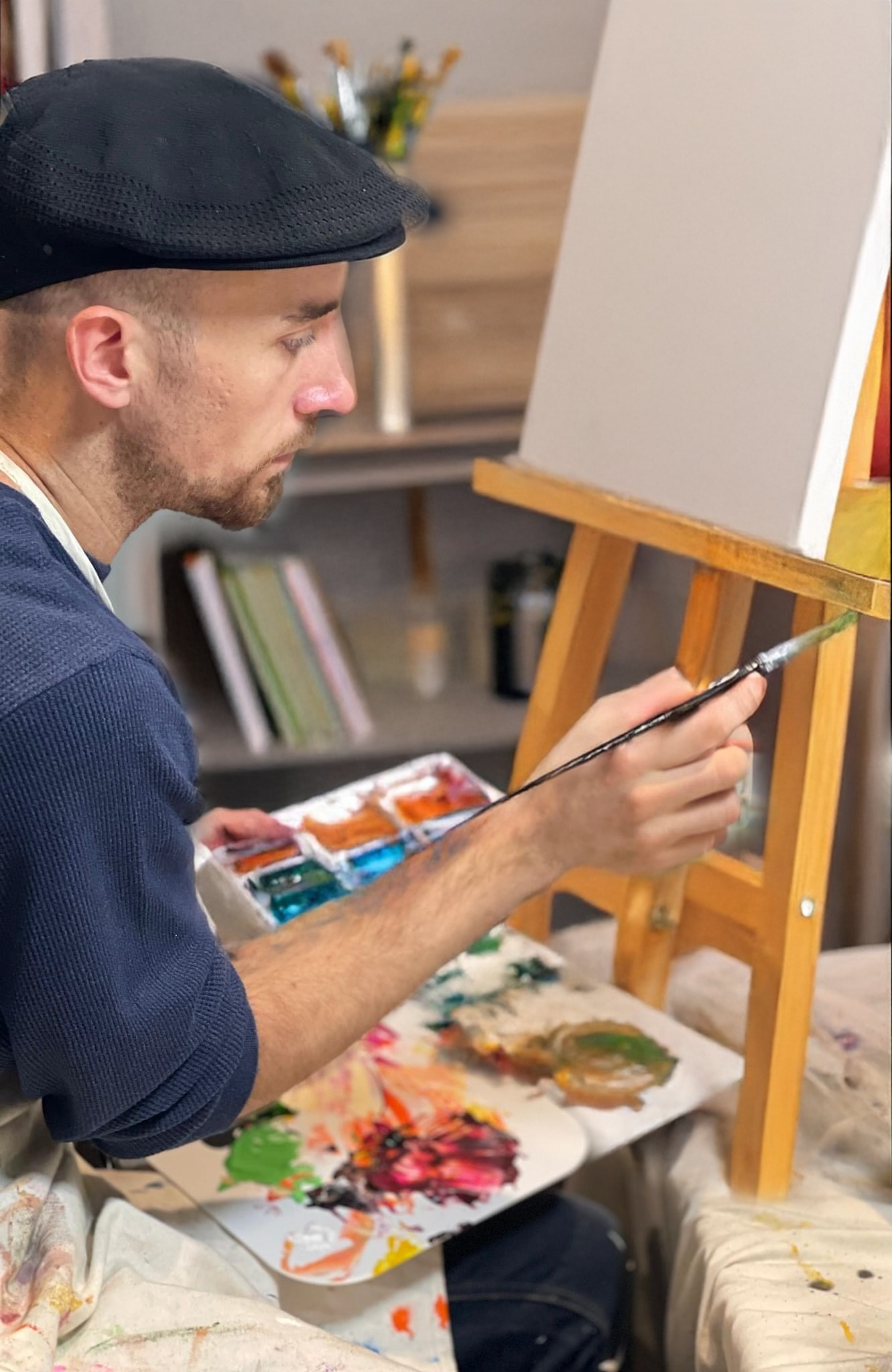 A man wearing a black cap is painting on a small canvas set on an easel. He holds a paintbrush and has a palette with various colors of paint. The workspace includes a cloth with paint splatters and a shelf with books and jars in the background.