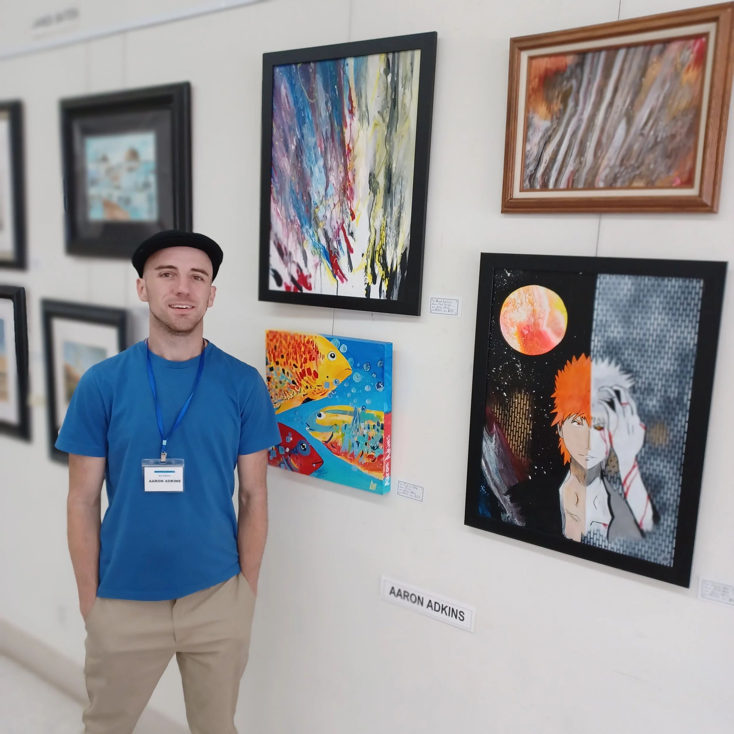 A man standing in front of abstract and colorful artwork at an art gallery, with some paintings featuring aquatic themes, vibrant colors, and a mix of abstract and portrait styles.