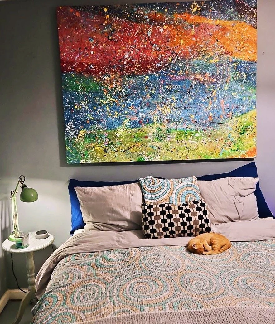 A bedroom with a large colorful abstract painting above the bed, pillows on the bed including patterned ones, a small orange kitten curled up and sleeping on the quilt, and a white nightstand with a green desk lamp, a candle, and a small speaker.