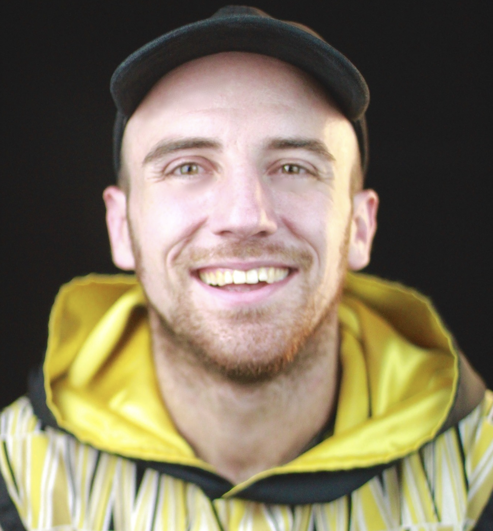 A smiling young man with a beard, wearing a black cap and a yellow jacket with a striped pattern.