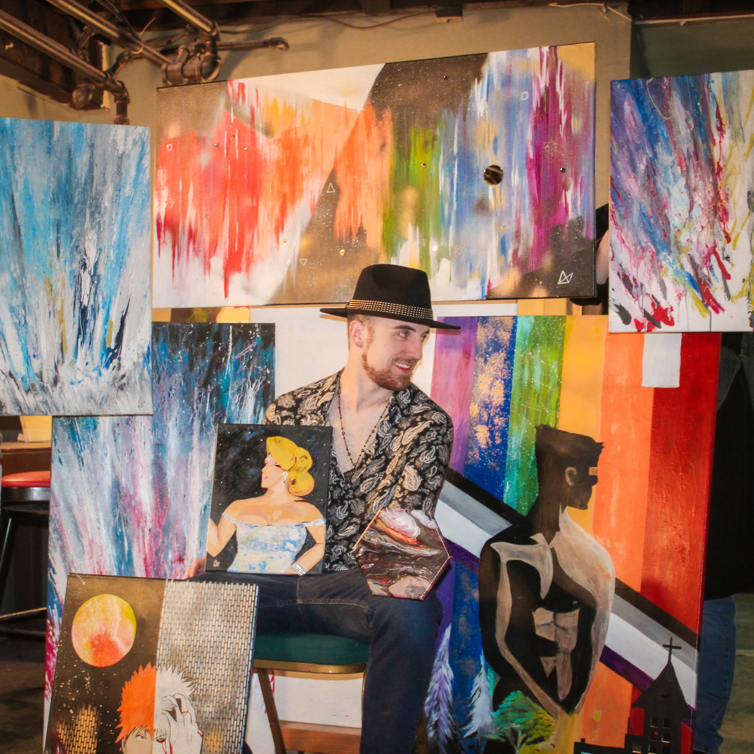 A man with a beard, wearing a black hat and patterned shirt, sitting among colorful abstract paintings at an art exhibition.