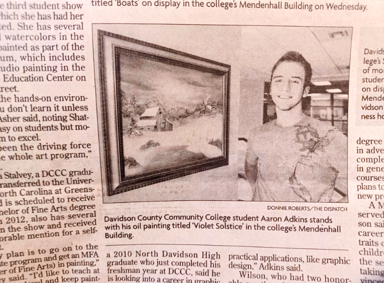 A young man standing next to a framed oil painting of a winter landscape with snow, trees, and a barn, in an indoor exhibition space.