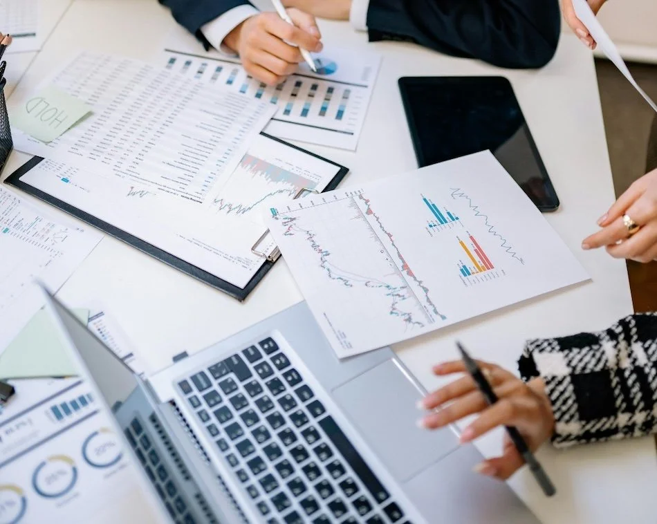 People analyzing financial charts and data reports on a desk with a laptop, tablet, and pens.