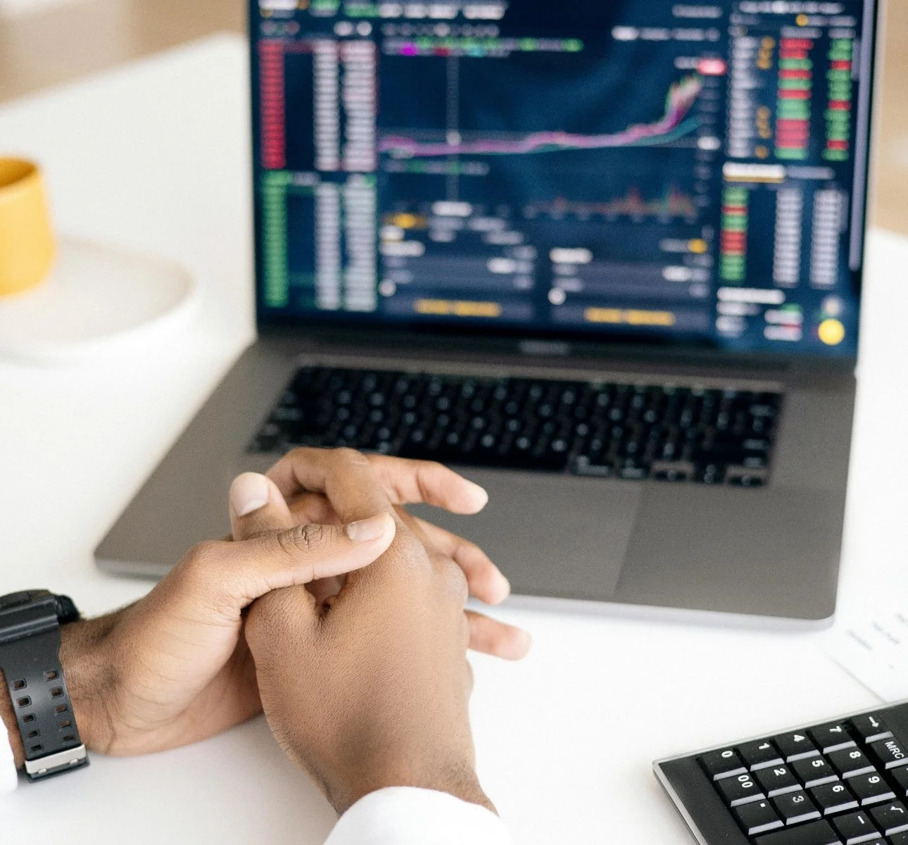 A person with dark skin and a black wristwatch sitting at a desk with a laptop displaying stock market charts and data, holding their hands clasped together.
