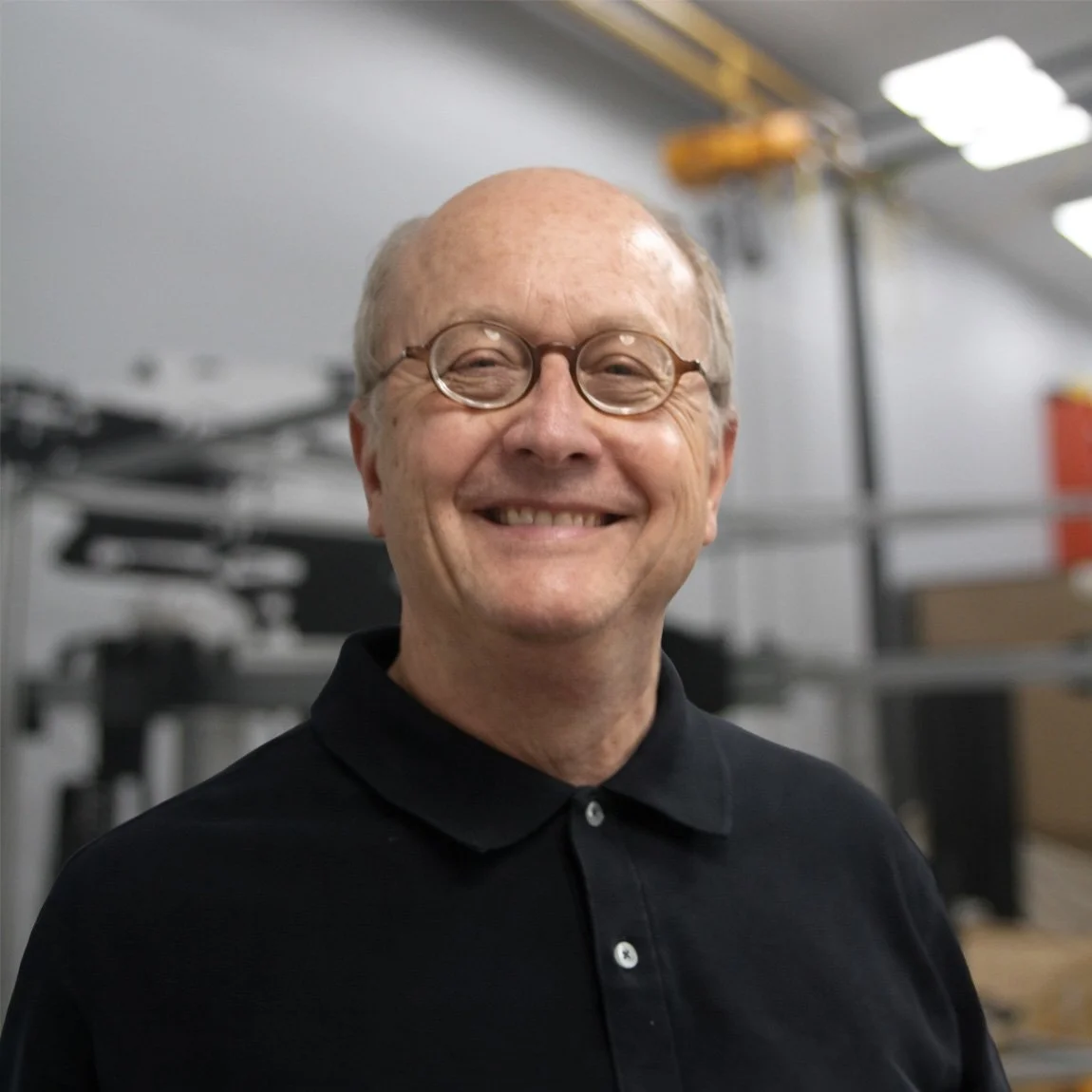A smiling man with glasses, wearing a black polo shirt, standing in a workshop or industrial setting with tools and equipment in the background.