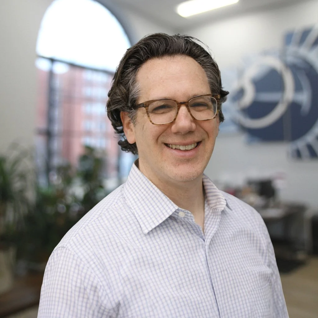 A smiling man with curly dark hair, wearing glasses and a light checkered shirt, standing in an office with large windows and a blurred background.