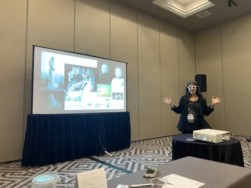 A woman giving a presentation in a conference room with a large screen showing people and a computer.