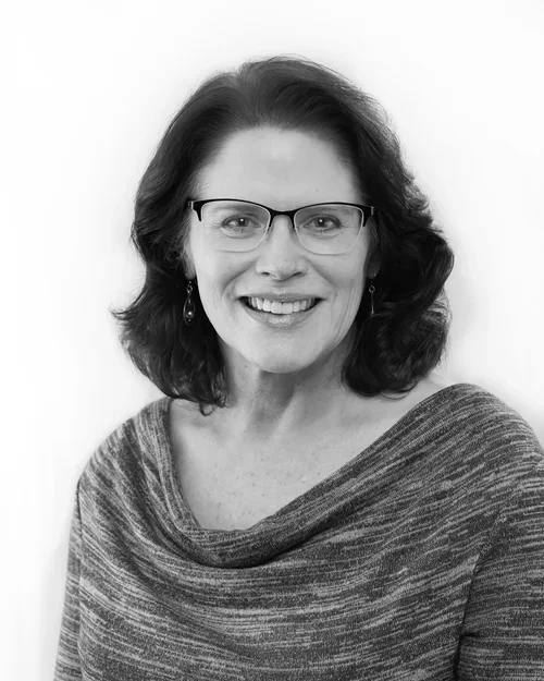 Black and white portrait of a woman with shoulder-length wavy hair, wearing glasses and a textured top, smiling at the camera.