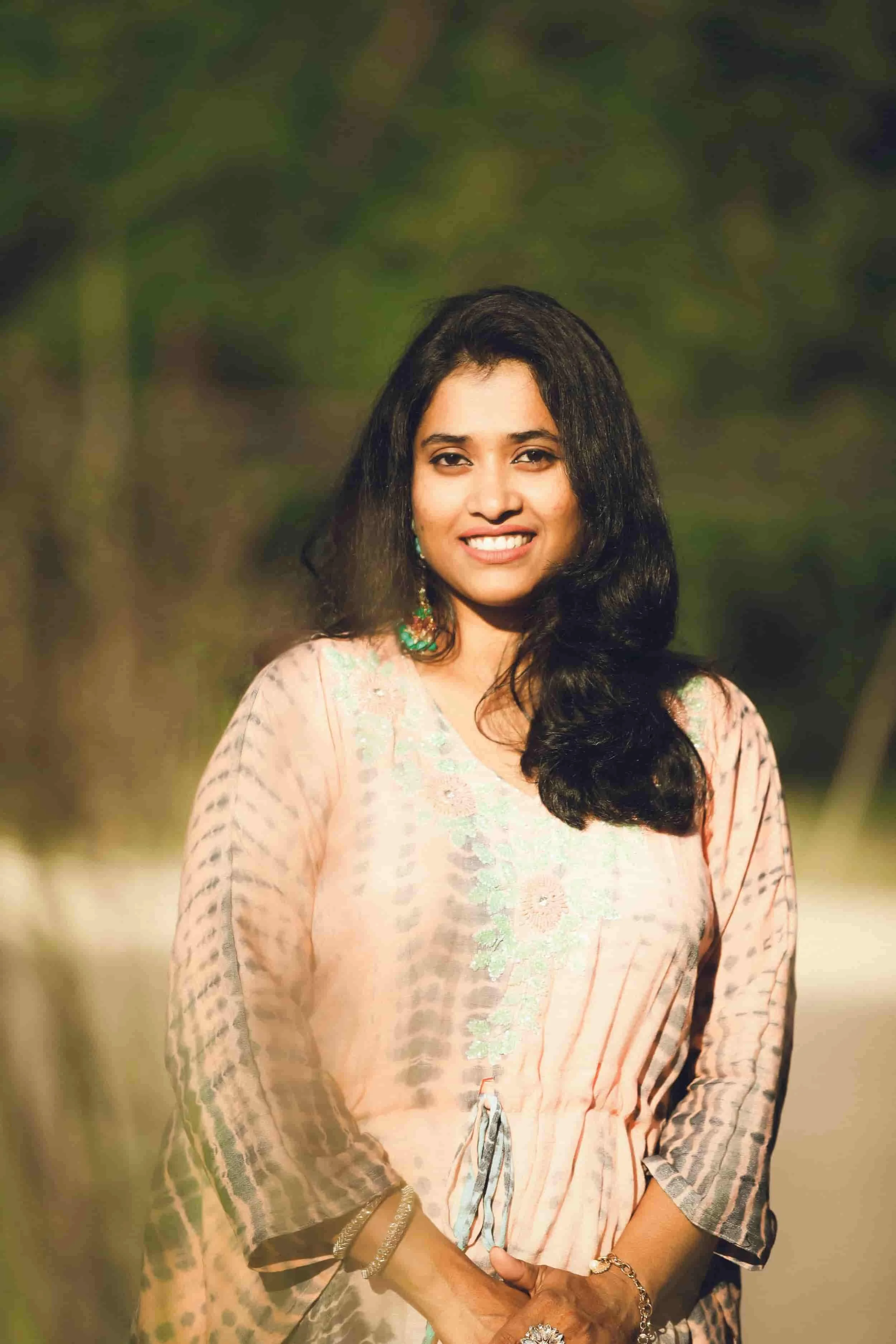 A woman with long black hair wearing a pink traditional dress with floral embroidery, standing outdoors against a blurred background of trees and greenery.