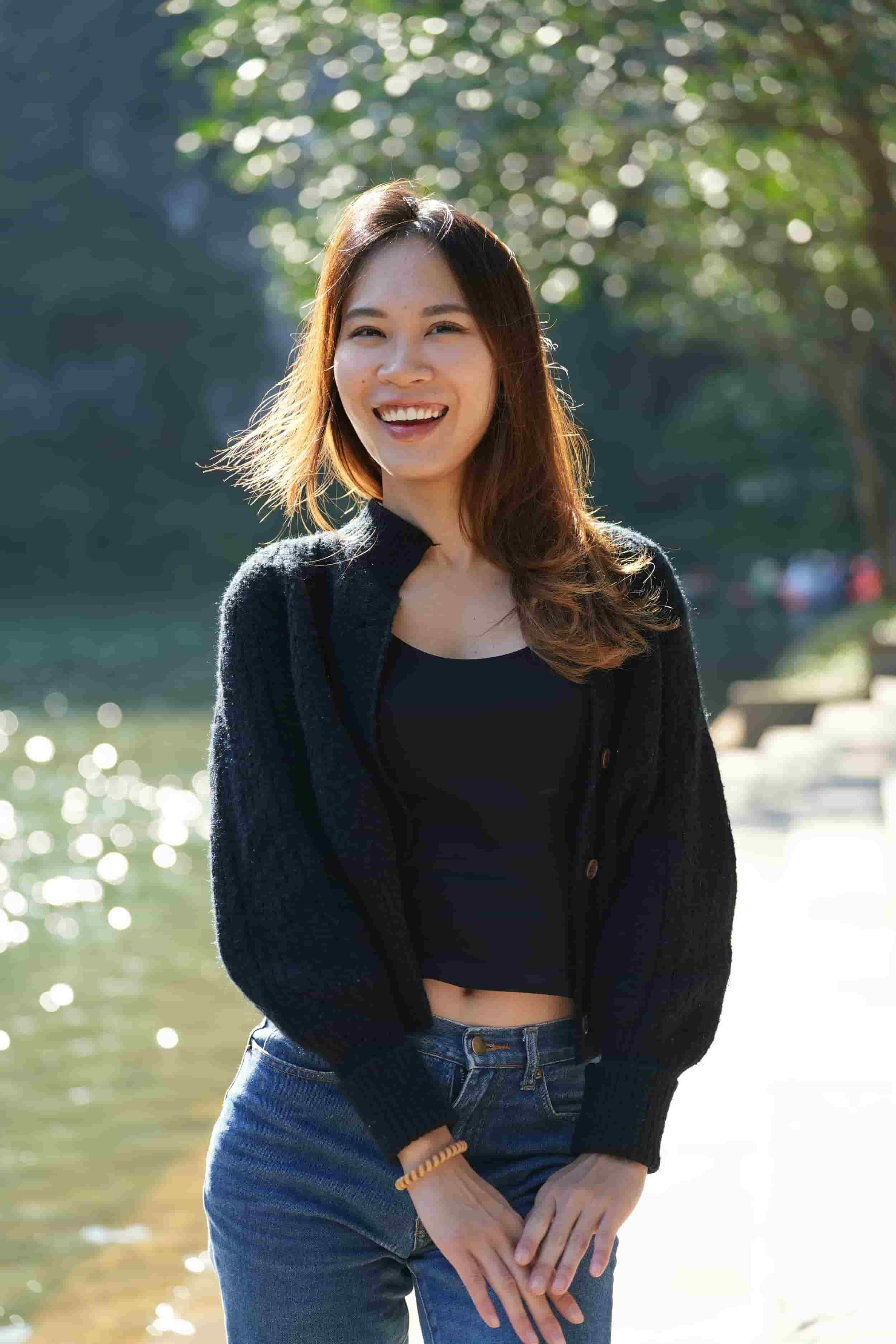 A young woman with shoulder-length brown hair smiling outdoors near a body of water on a sunny day.