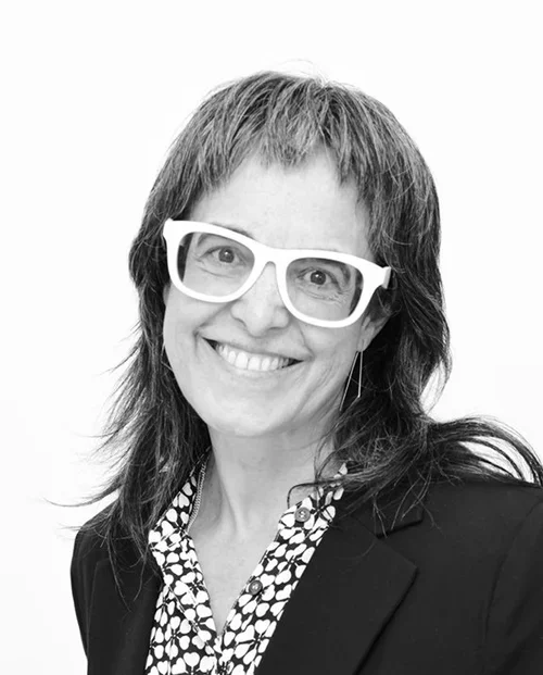 Black and white portrait of a woman with medium-length hair, white glasses, smiling, wearing a blazer over a patterned blouse.