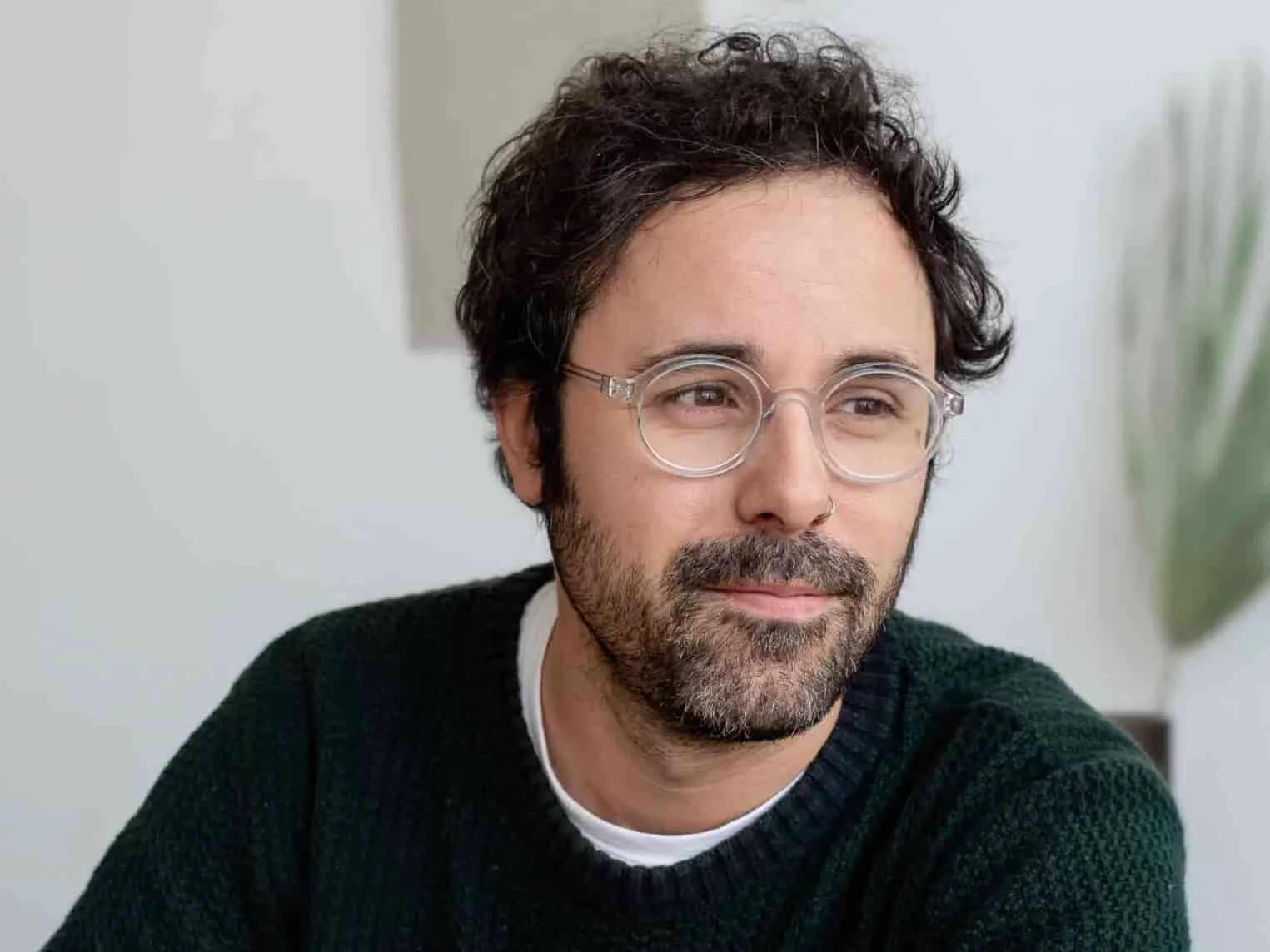 A man with glasses and a nose piercing, wearing a black sweater, looks to the side with a slight smile. A blurred plant is in the background.