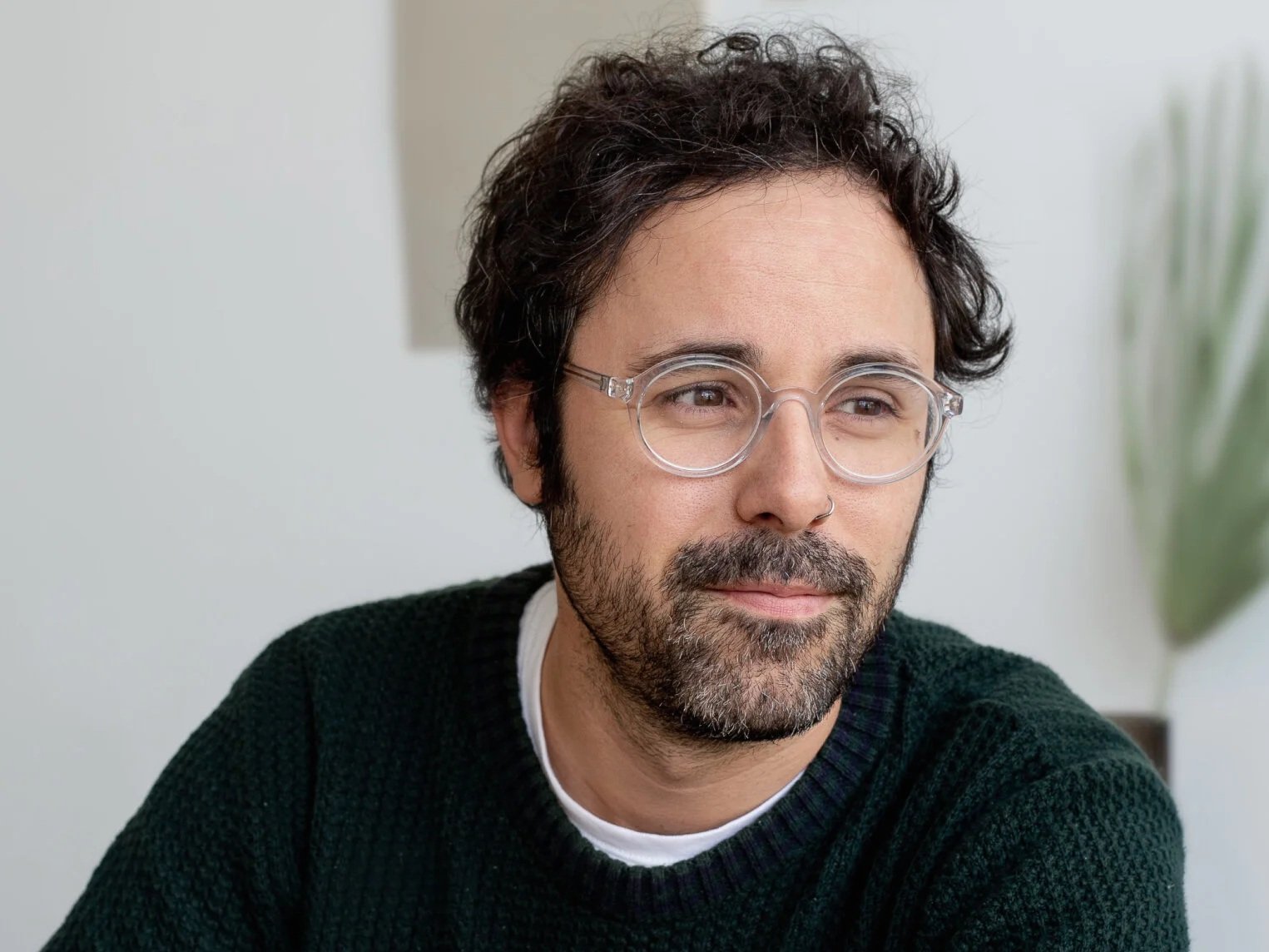 A man with glasses and a nose piercing, wearing a black sweater, looks to the side with a slight smile. A blurred plant is in the background.