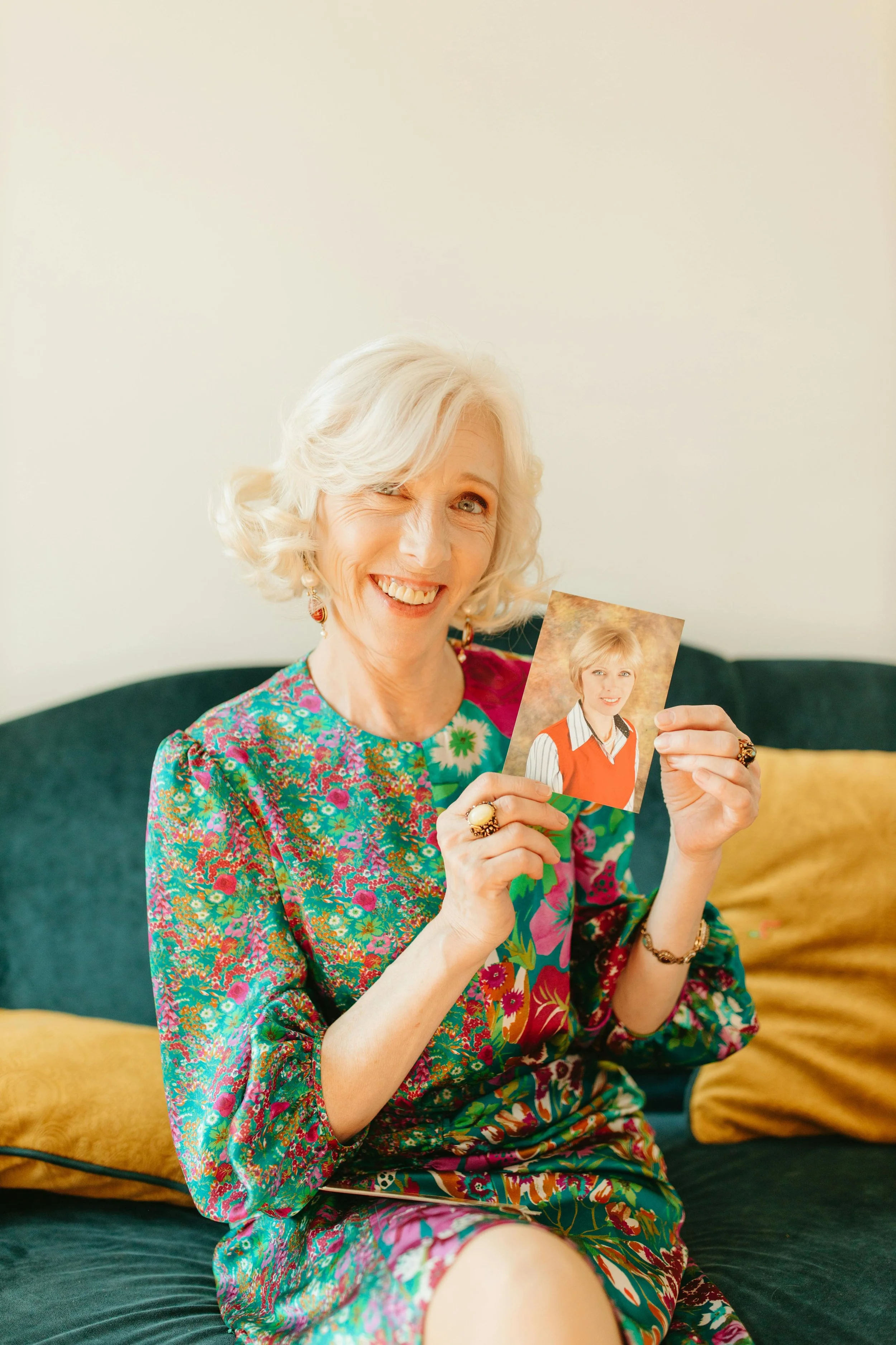An elderly woman with curly white hair smiling and holding a photograph of herself when she was younger, sitting on a dark green sofa with yellow pillows.