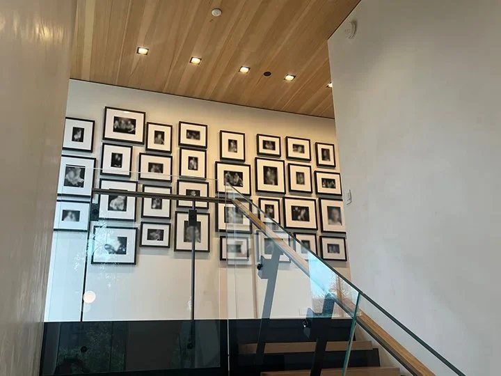Interior view of a staircase with a glass railing, adjacent to a white wall decorated with numerous black-and-white framed photographs arranged in a grid pattern.