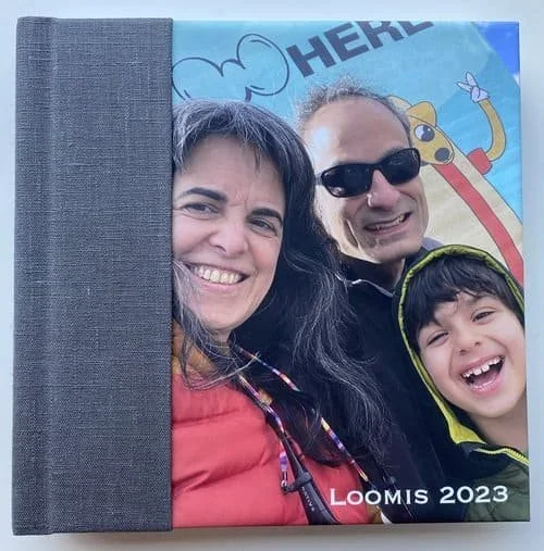 A smiling woman, man wearing sunglasses, and young boy in a yellow hoodie taking a selfie with a colorful cartoon background in front of a book or photo album.