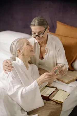 An elderly woman with white hair and a caregiver with glasses sitting together, looking at each other warmly in a bedroom.