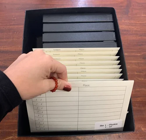 Hand holding a sheet of index cards with 'Subject' and 'Place' columns inside a black box on a wooden surface.
