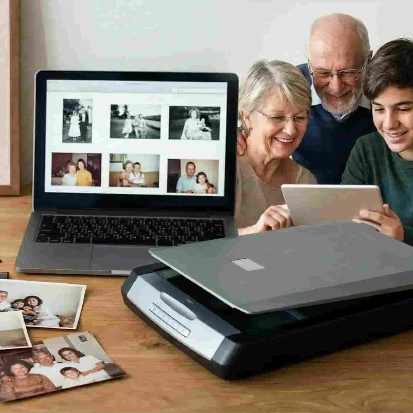 Three generations of family members smiling while looking at a tablet together, with a laptop displaying old photographs and printed printed photos on the table.