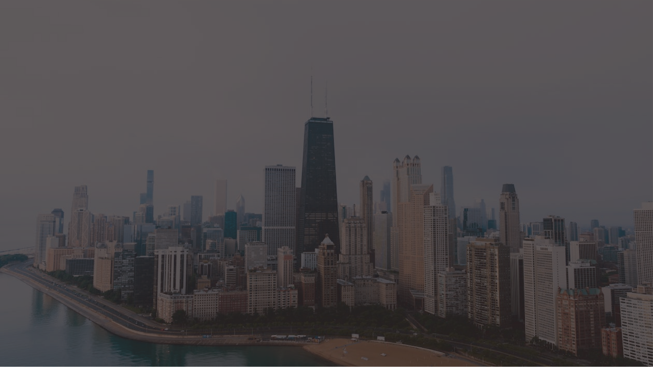 Aerial view of a city skyline with tall skyscrapers including the John Hancock Center, along a river with a beach in the foreground, under an overcast sky.