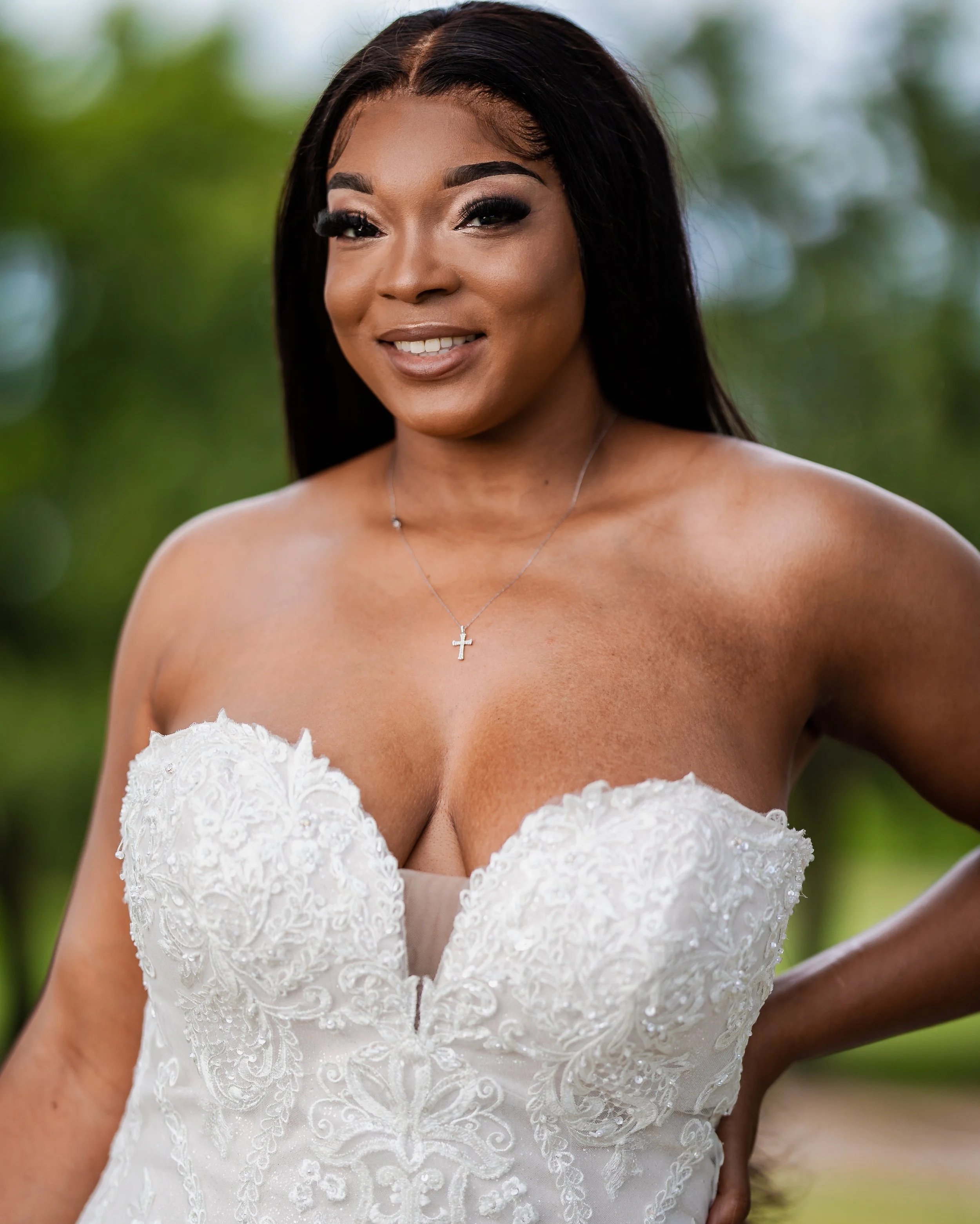 A woman in a strapless white wedding dress with lace embroidery, standing outdoors with a blurred green background.