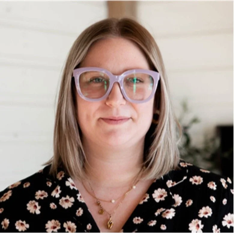 A woman with shoulder-length gray hair wearing large lavender glasses, a black floral top, and layered necklaces, standing indoors with a white wall and some plants in the background.