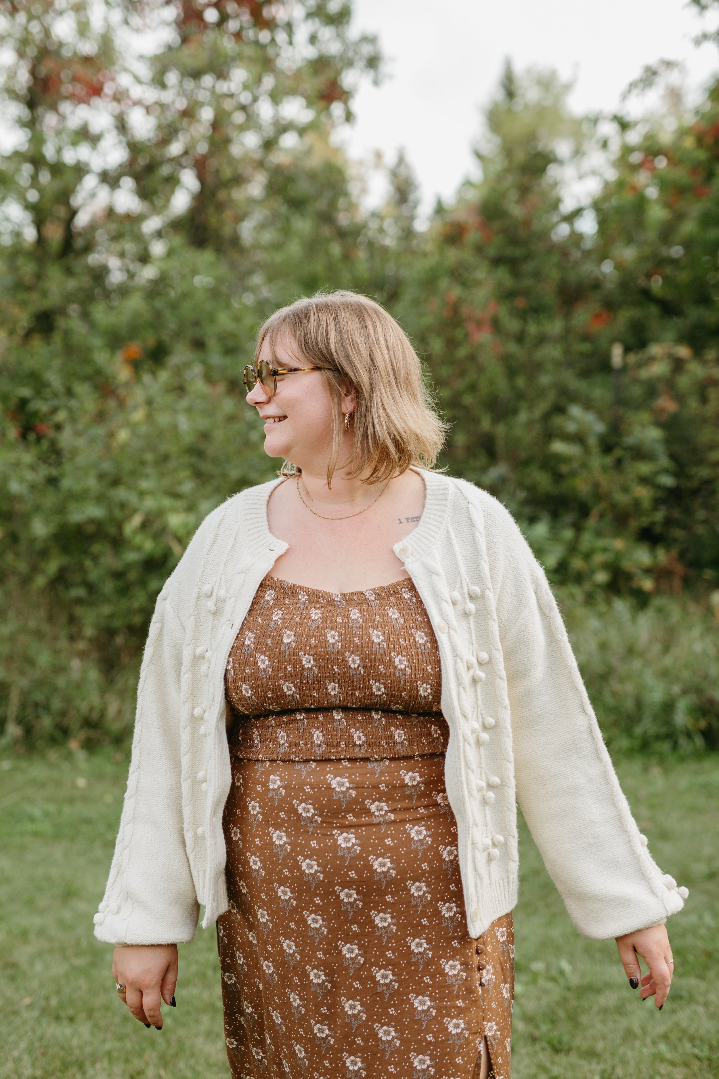 A woman with shoulder-length light brown hair, wearing sunglasses, a white cardigan, a brown floral dress, smiles as she stands outdoors in a grassy area with trees in the background.