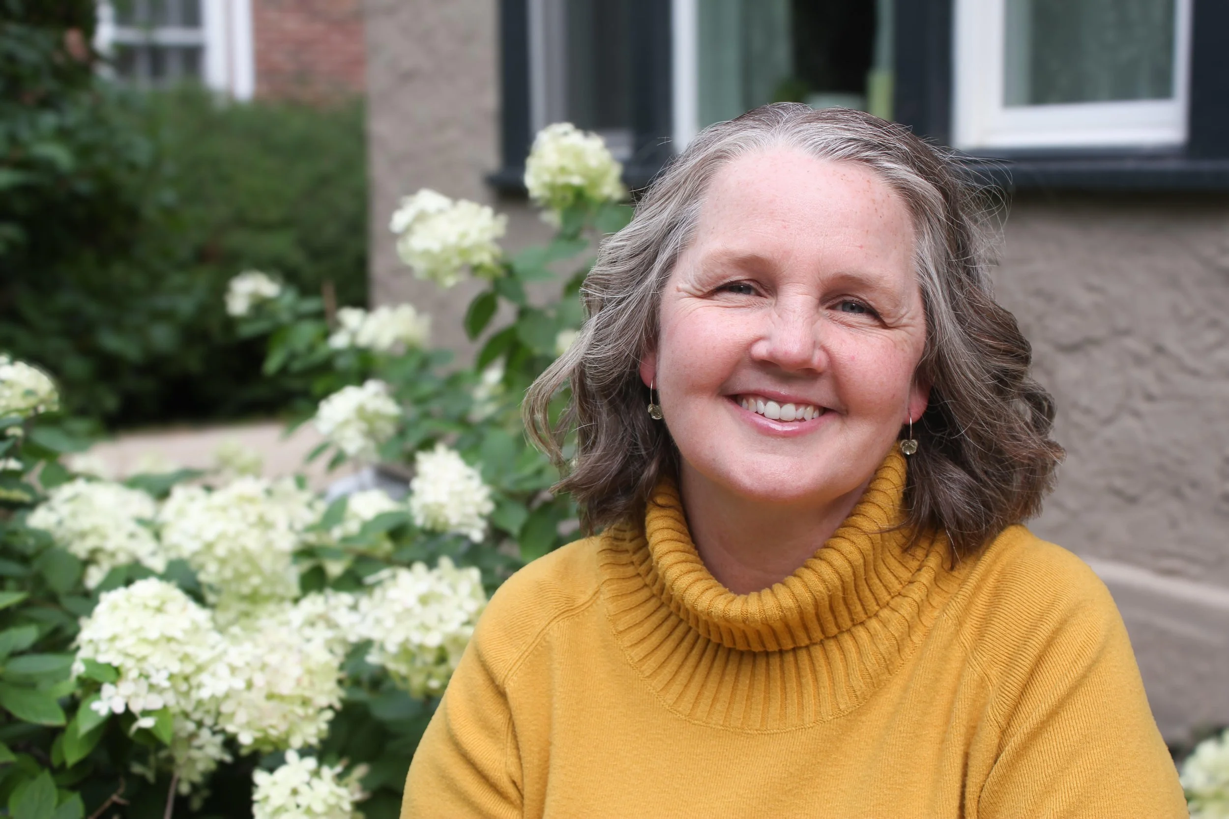A smiling woman with shoulder-length wavy gray hair wearing a yellow turtleneck sweater, standing outdoors near white hydrangea flowers and a house with a gray brick exterior.