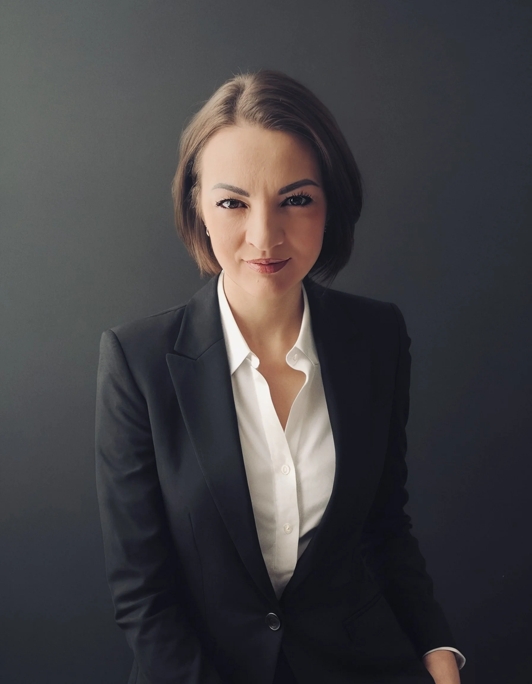 A woman with short brown hair, wearing a black blazer and white blouse, stands against a dark grey background, looking directly at the camera with a confident expression.