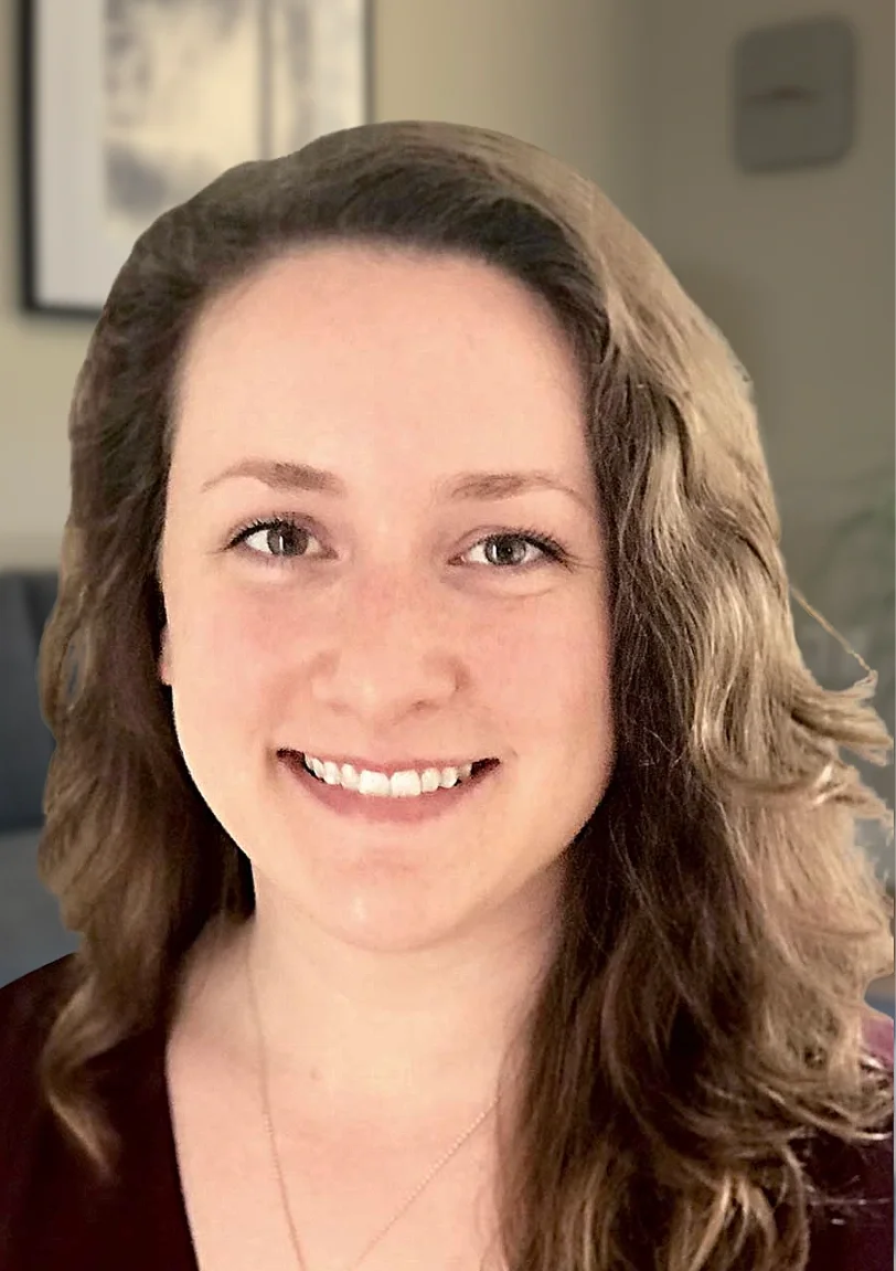 A smiling woman with long, wavy brown hair, light skin, and light-colored eyes indoors with blurred background.