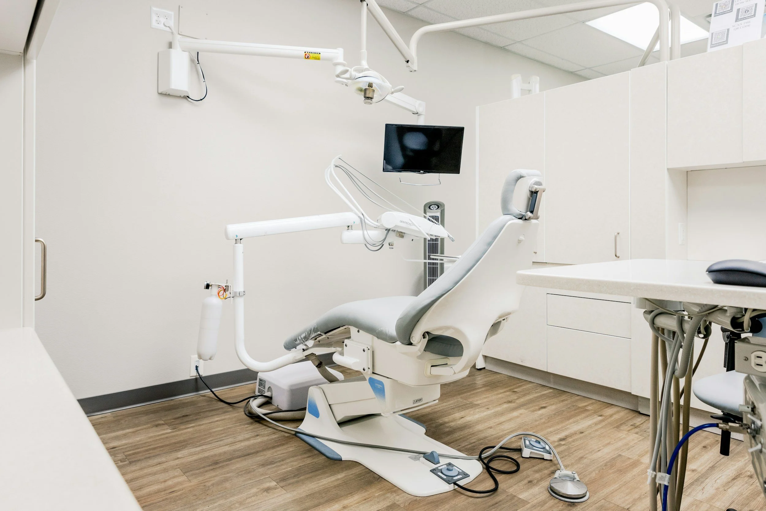 A dental examination room with a dental chair, computer monitor, and various dental equipment on the wall and nearby counter.
