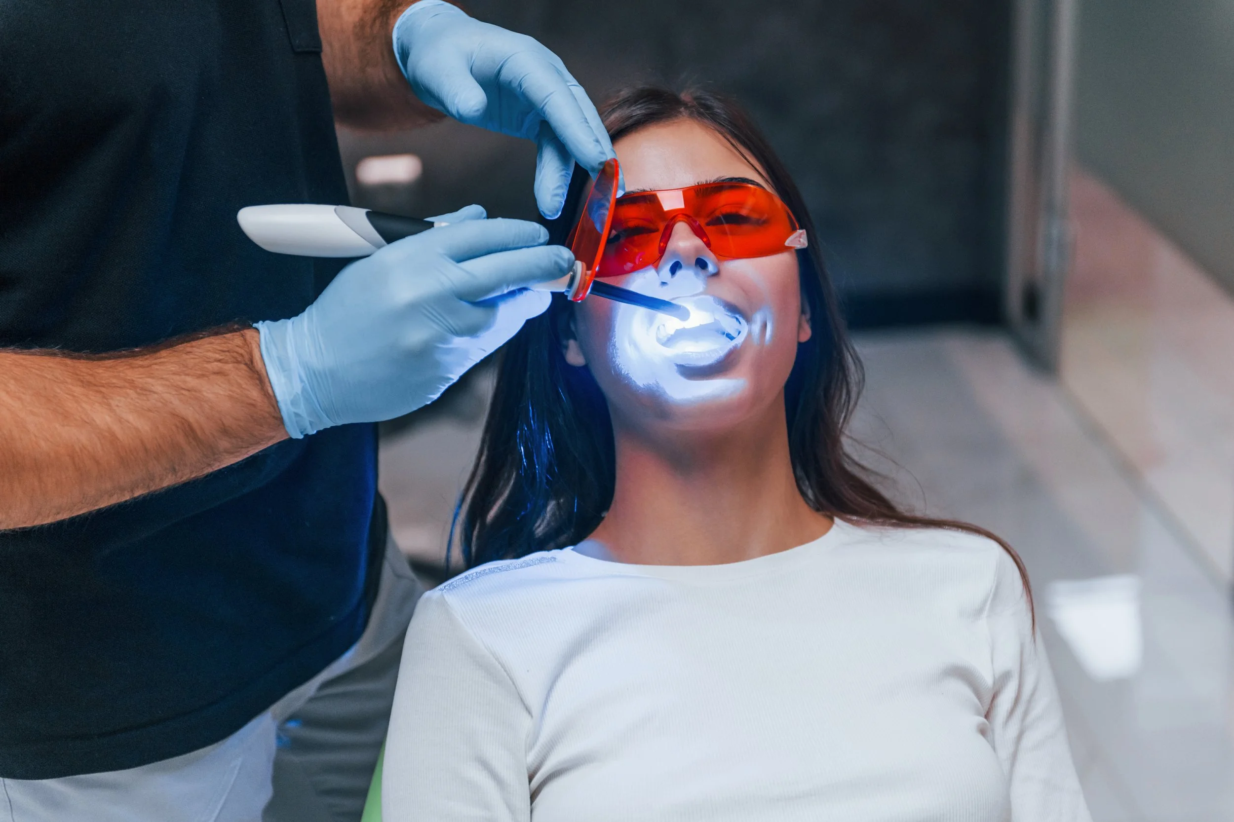 A woman receiving dental treatment with a handheld curing light, wearing protective orange glasses, while a dentist in blue gloves works on her mouth in a modern dental clinic.