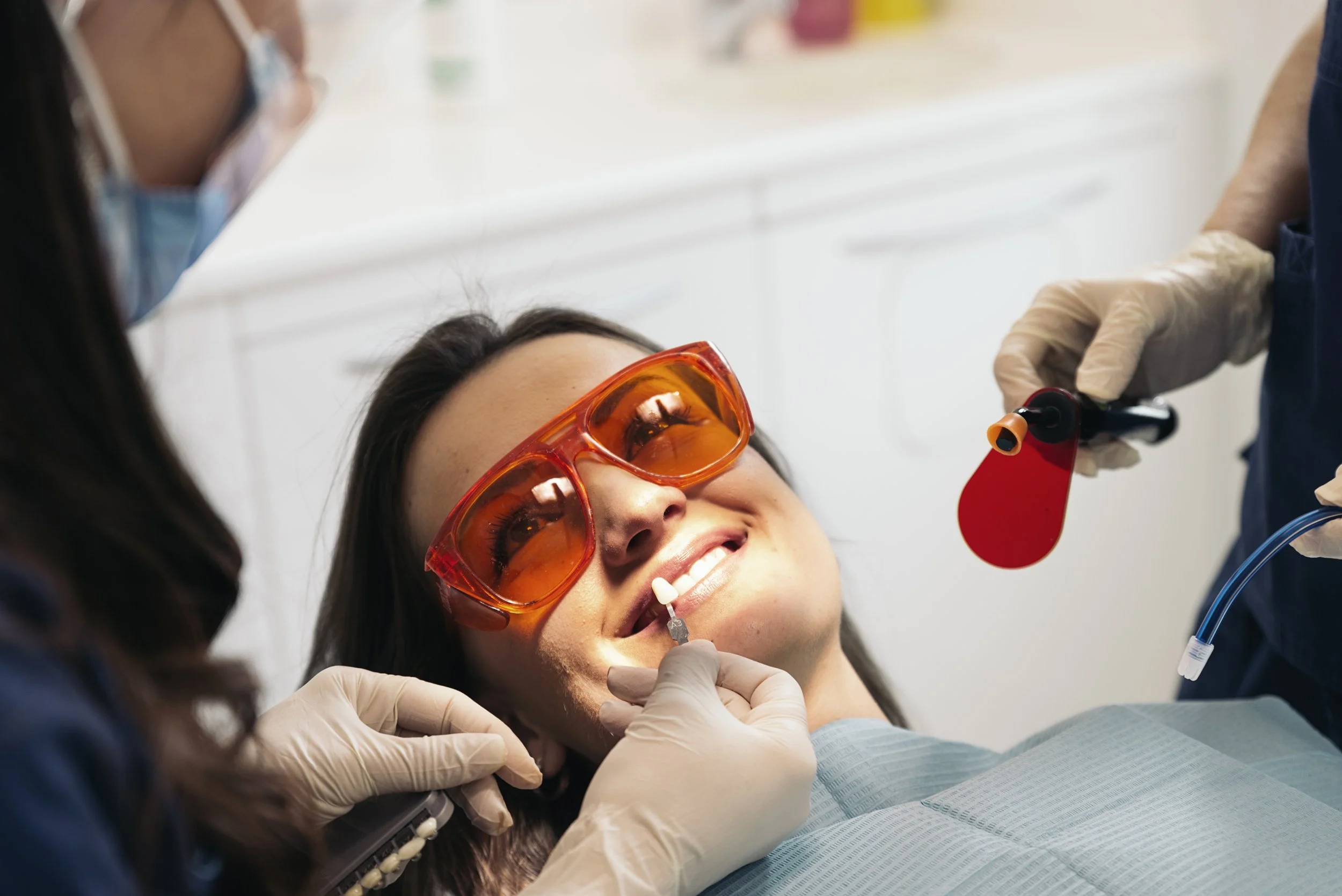 A woman lying on a dental chair, smiling during a dental procedure, with protective orange glasses. Dental professionals wearing gloves are administering treatment using dental tools.
