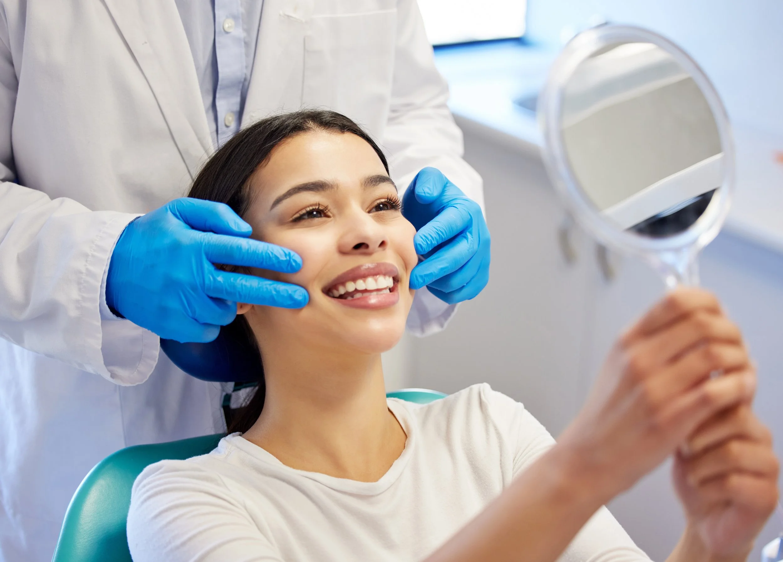 A woman sitting in a dental chair smiling while a dental professional, wearing blue gloves and a white coat, holds her face and looks at her reflection in a mirror.