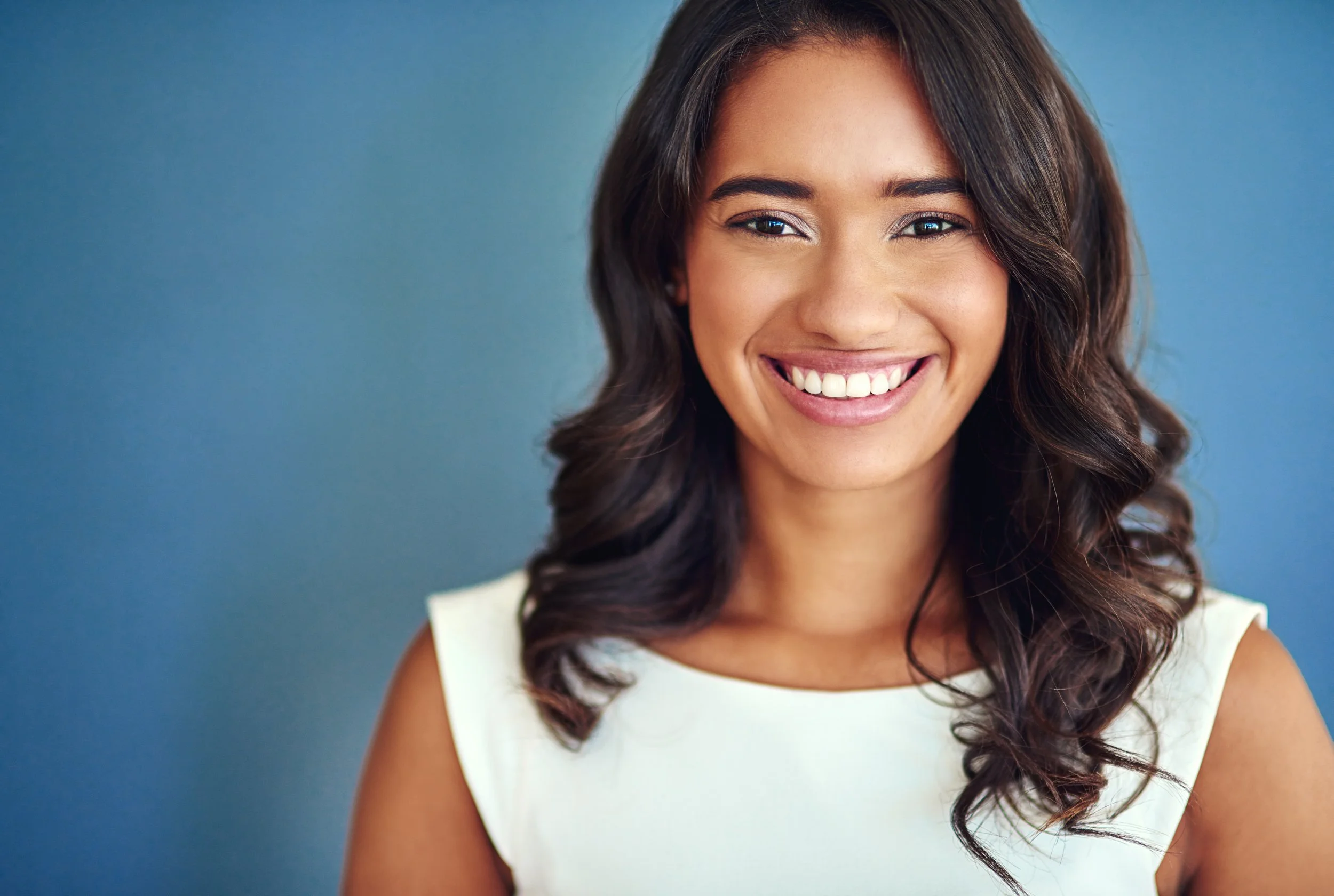 A smiling woman with long, dark, wavy hair wearing a sleeveless white top, standing against a blue background.