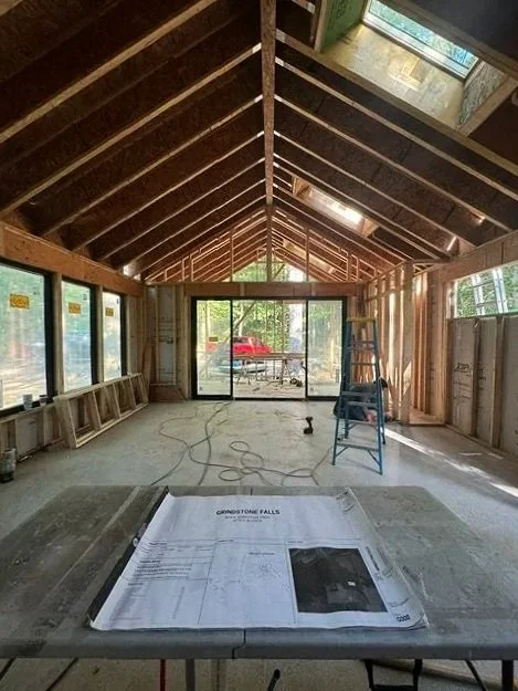 Unfinished room under construction with exposed wooden framing, large windows, a sliding glass door, a ladder, and construction plans on a table.