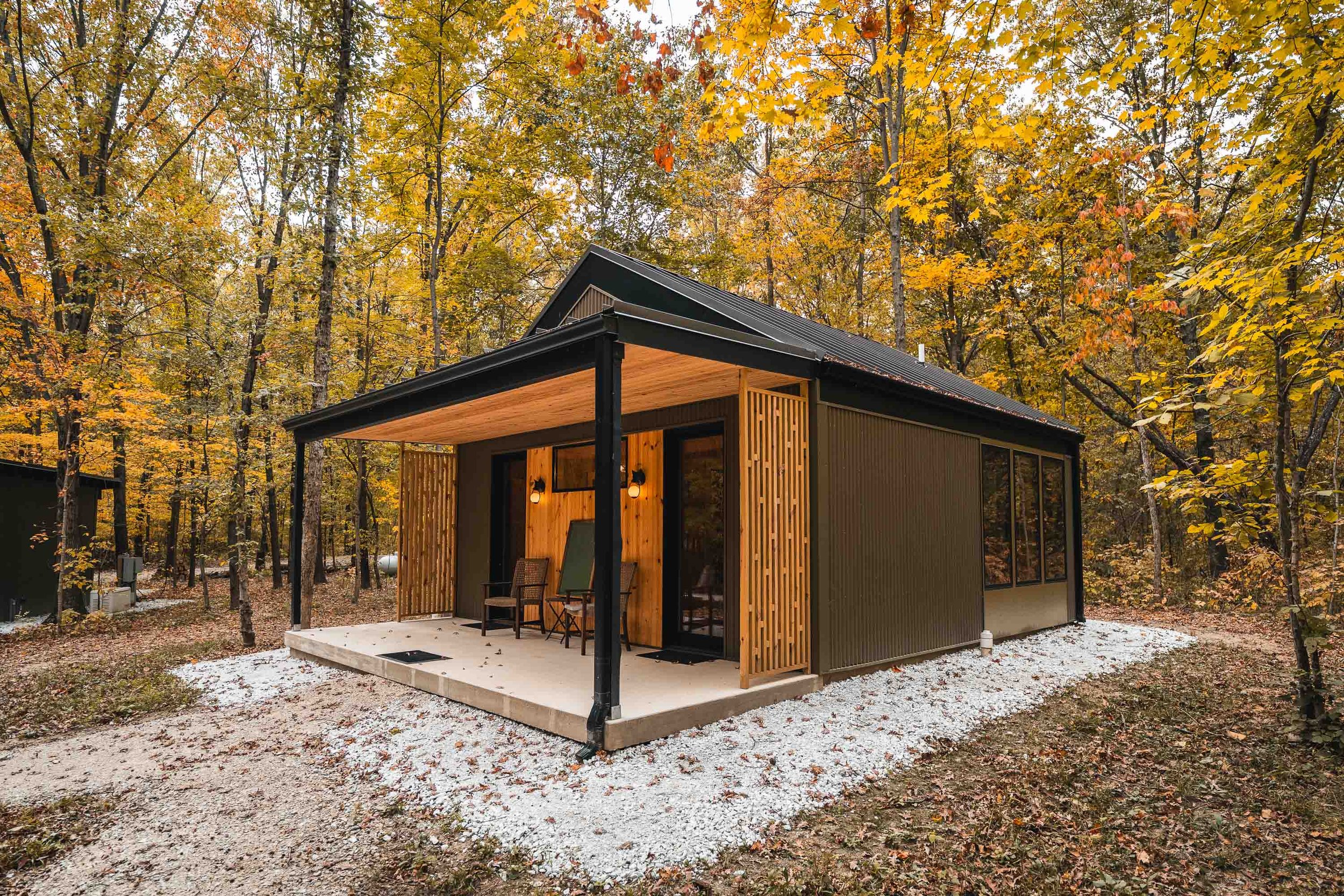 A small modern house with a porch, surrounded by trees with autumn foliage, and a gravel path in front.