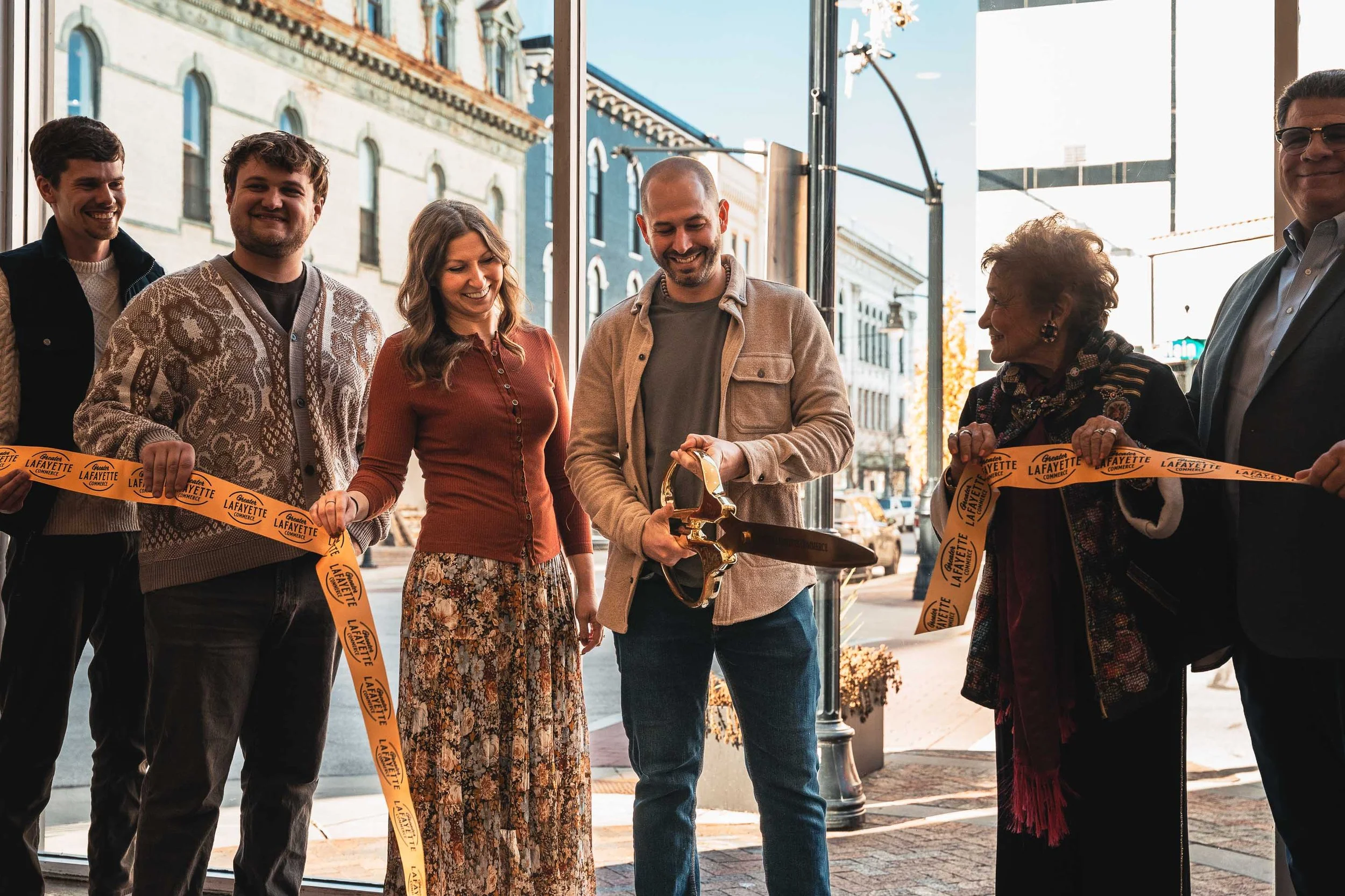 People attending a ribbon-cutting ceremony outside a storefront, holding a yellow ribbon with the text 'Lafayette.' A man in the center is holding a large pair of scissors ready to cut the ribbon, with others smiling and watching.