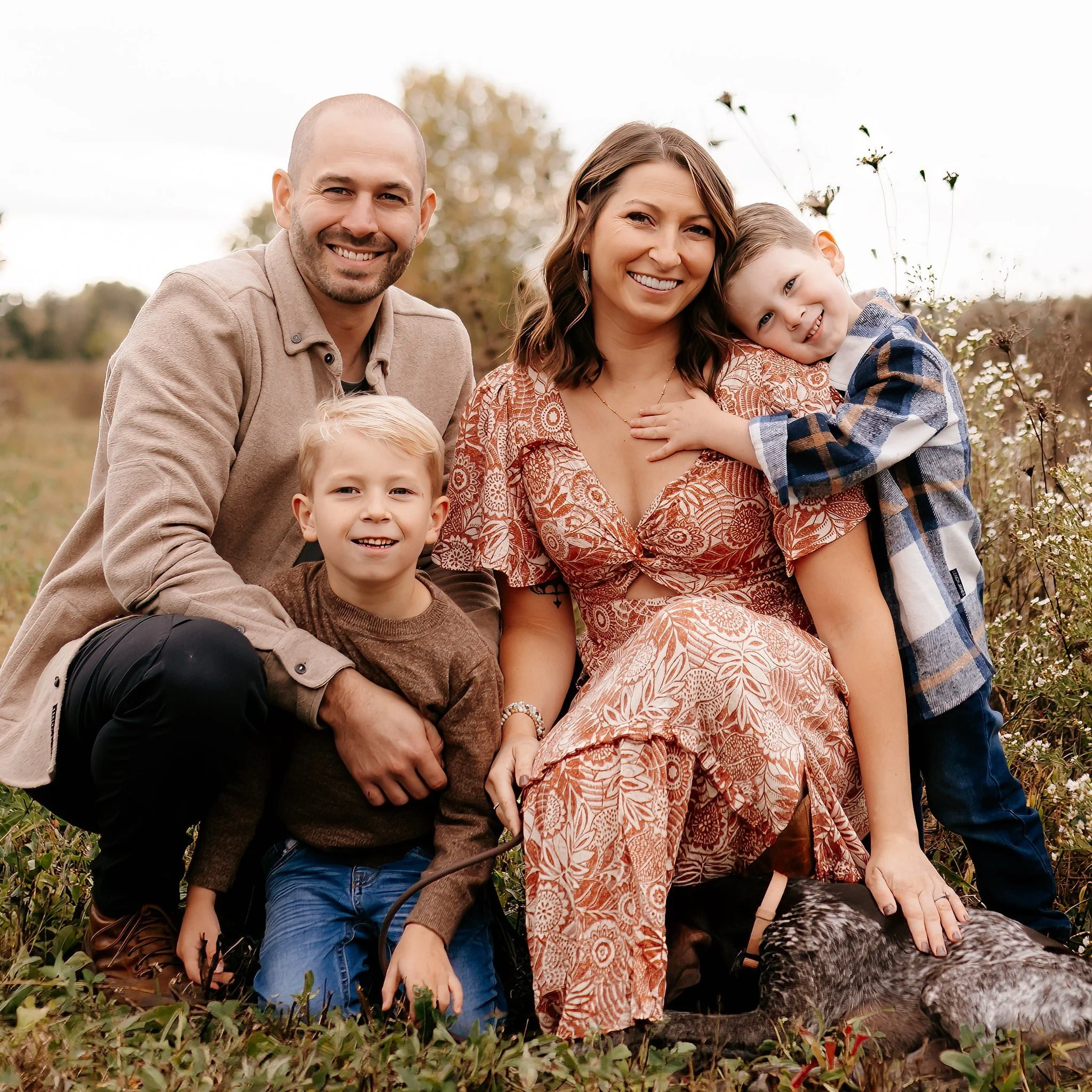 Family of five, including mother, father, two young boys, and a girl, posing outdoors in a field with a dog. Everyone is smiling.