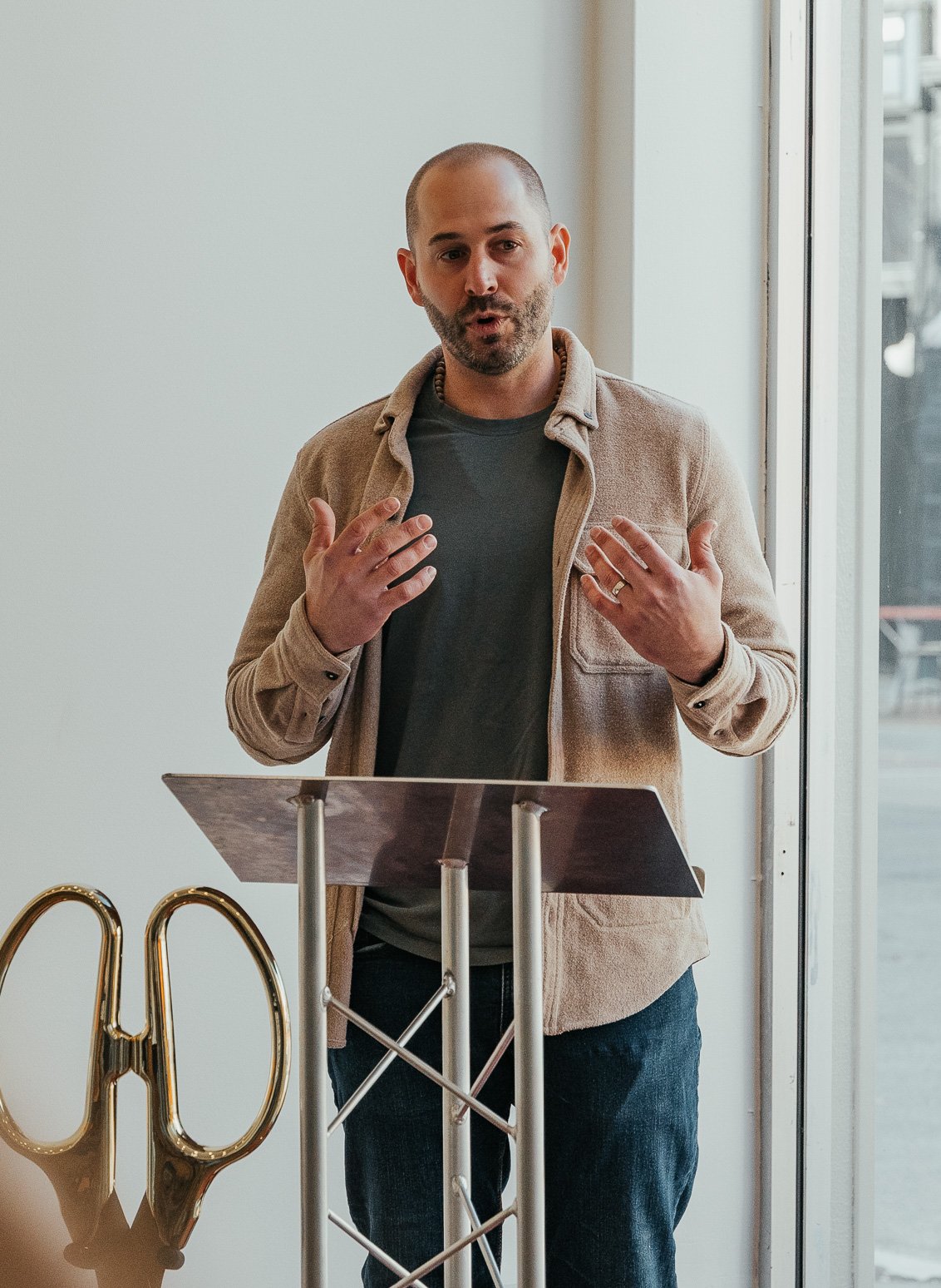 A man standing behind a podium with a laptop, speaking or presenting, with large gold scissors nearby, near a window.