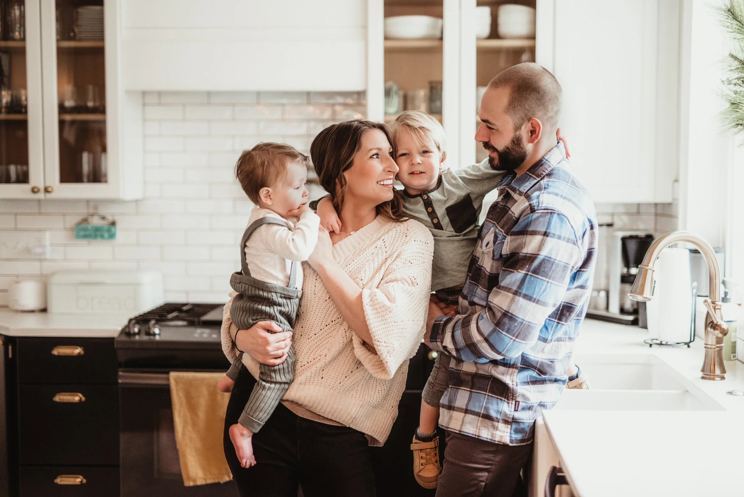 A smiling family of four with two young children in the kitchen, with the mother holding one child and the father holding the other, sharing a joyful moment.