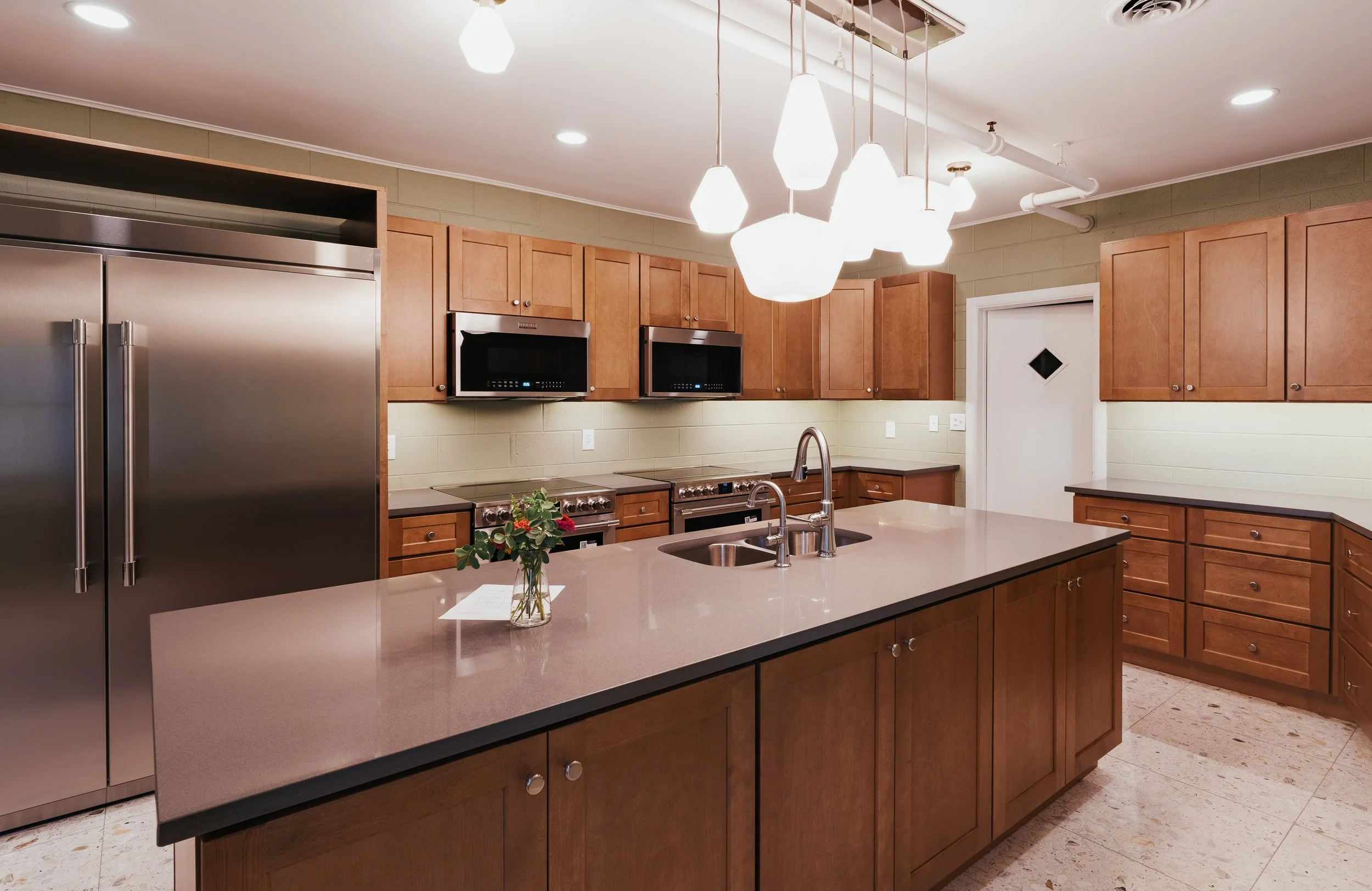 Modern kitchen with wooden cabinets, stainless steel refrigerator, and built-in microwave. An island with a sink and a small vase of flowers on top. Bright lighting from hanging pendant lights and ceiling lights.