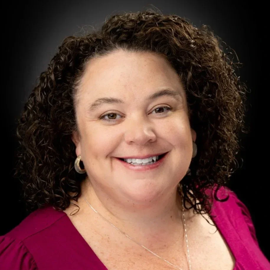A woman with curly brown hair wearing a magenta top and gold jewelry smiling at the camera