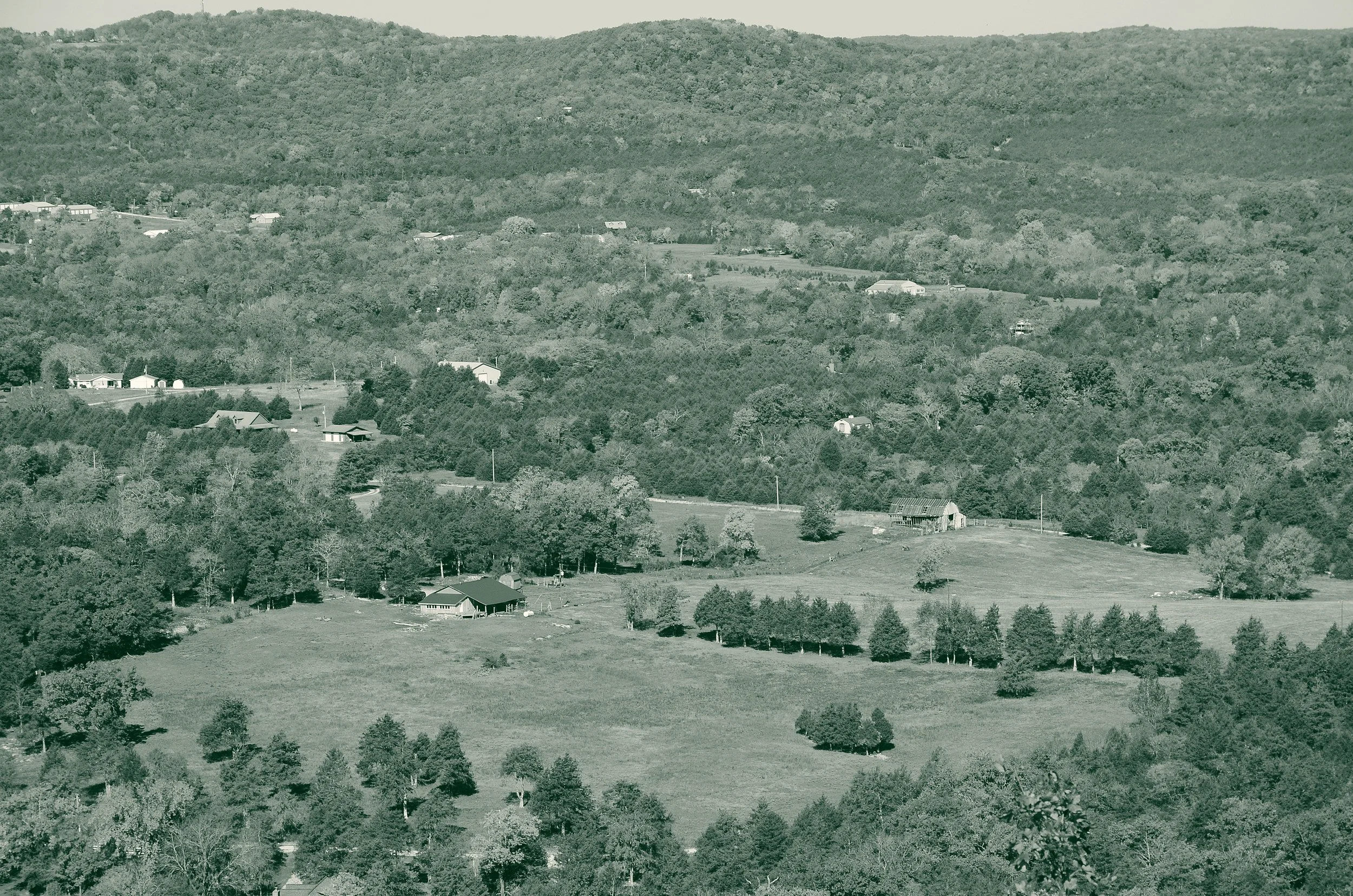 A farmhouse and barn in a White River valley in the Ozark Mountains of Arkansas, with pasture, tree lines and scattered rural homes across the wooded hills.