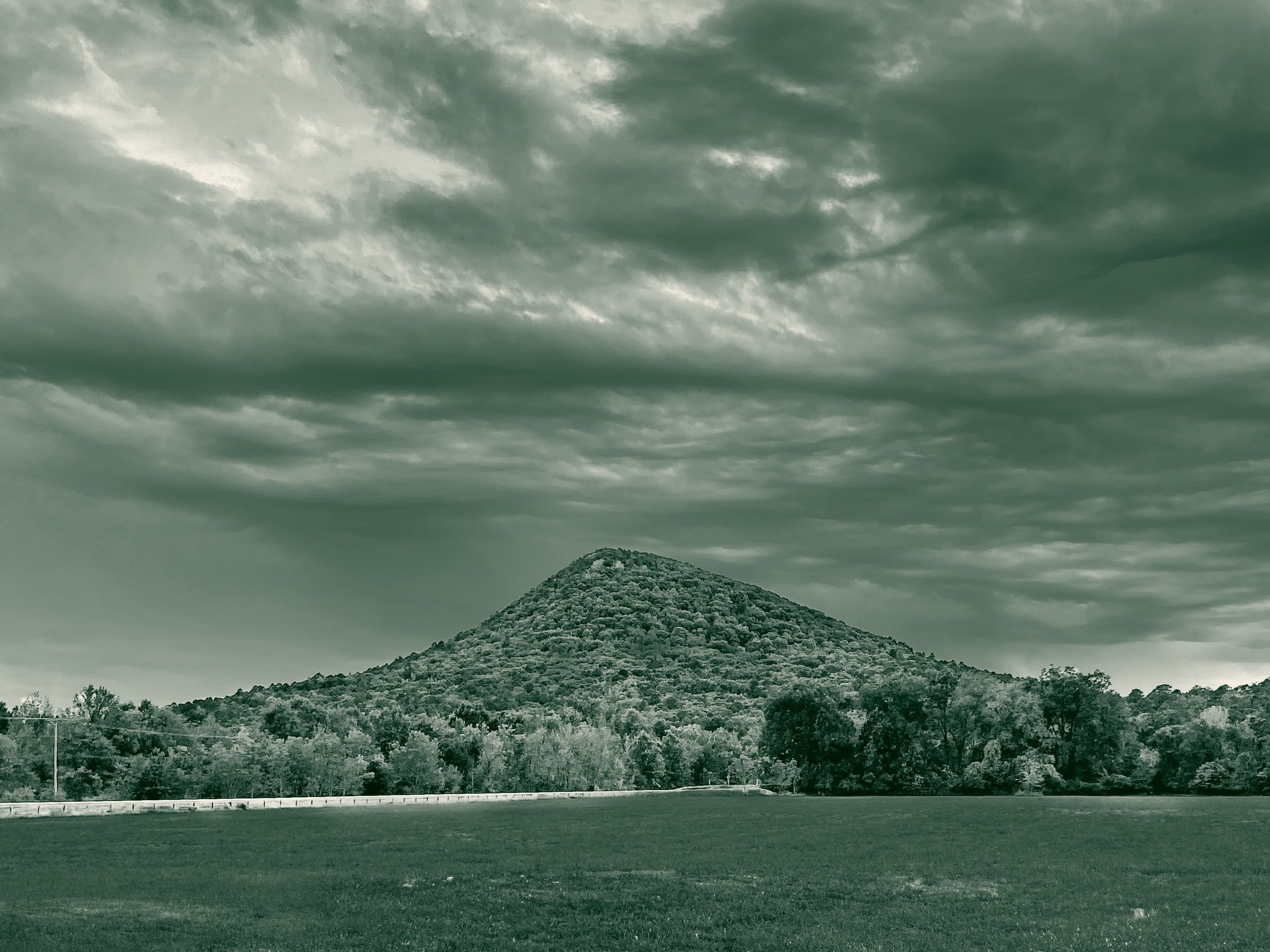 Pinnacle Mountain rises above a wooded treeline and an open field in Pulaski County, Arkansas, beneath a broad, heavily clouded sky.