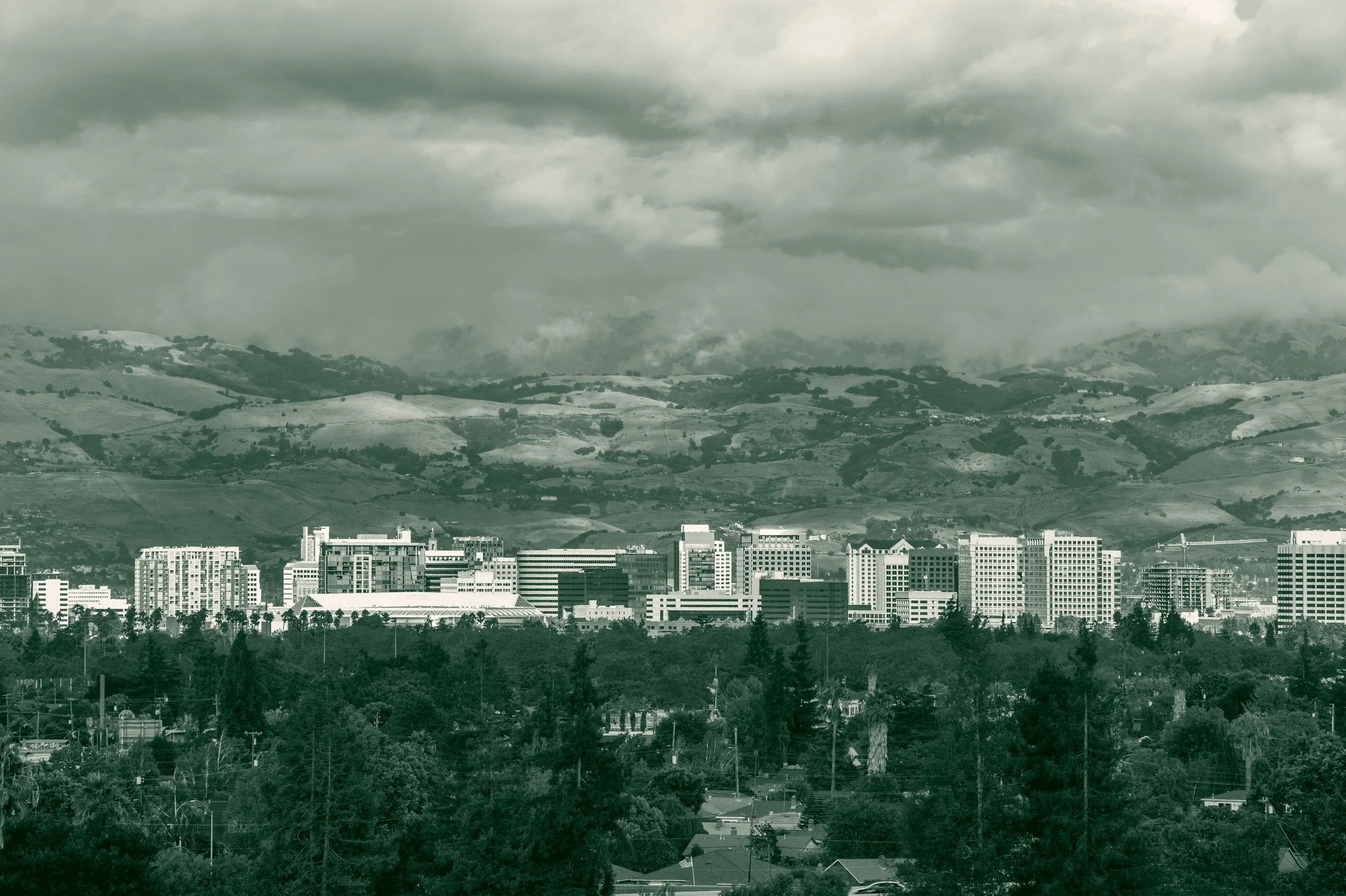 Downtown San Jose seen from across the valley, with residential tree canopy in the foreground and the hills of the Diablo Range behind under a clearing storm.
