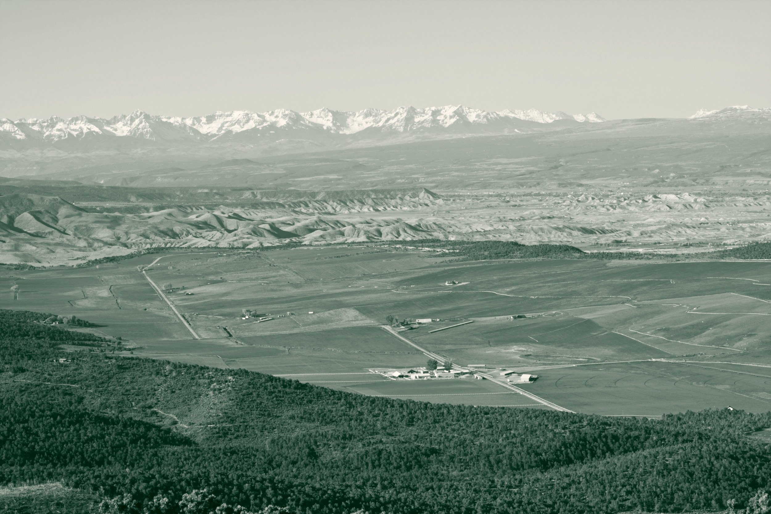 A wide view across the Gunnison valley in western Colorado, with irrigated farmland and forested hillsides below a snowcapped mountain range.