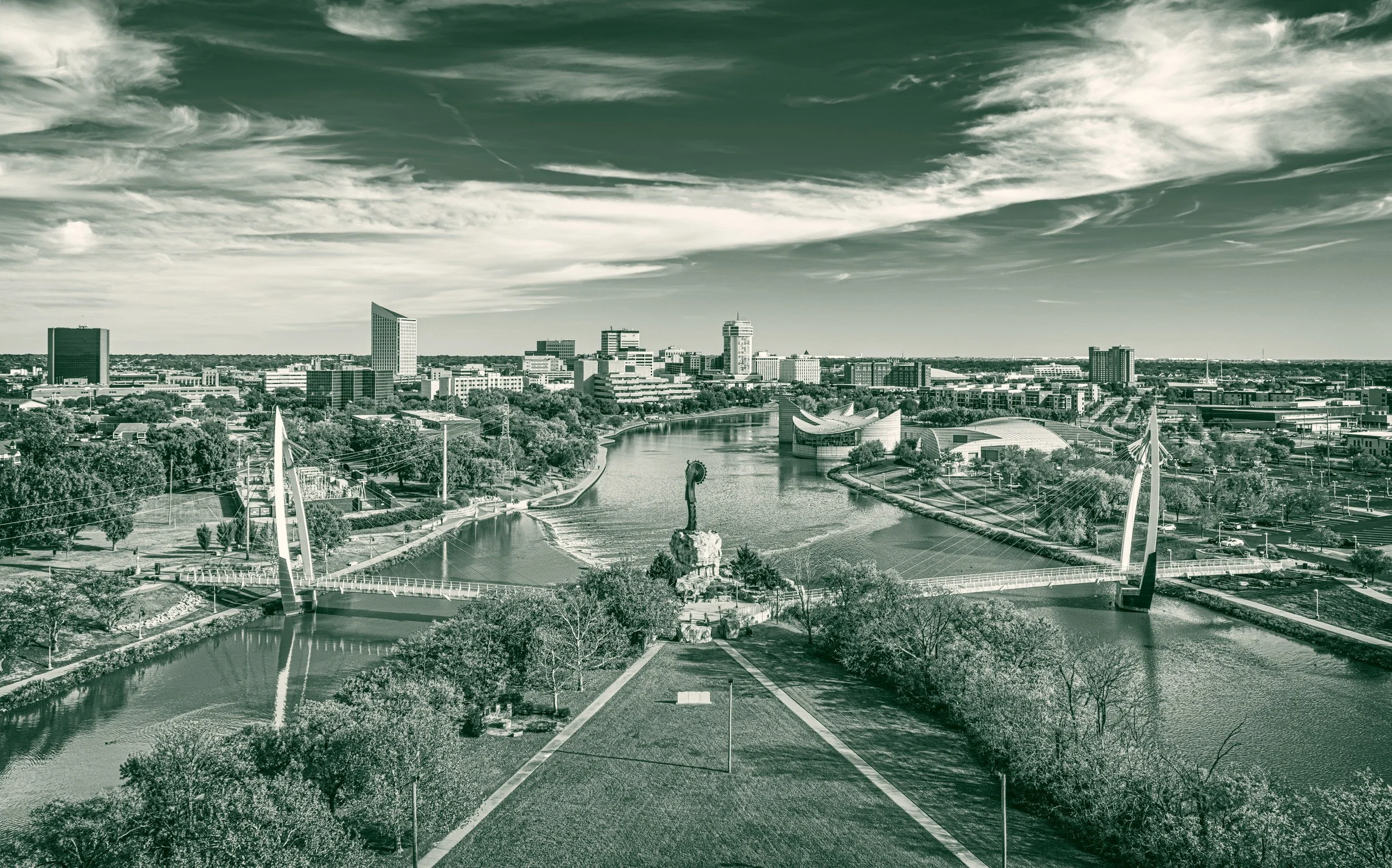 The Keeper of the Plains sculpture at the confluence of the Arkansas and Little Arkansas rivers, with two pedestrian bridges and the downtown Wichita skyline behind.
