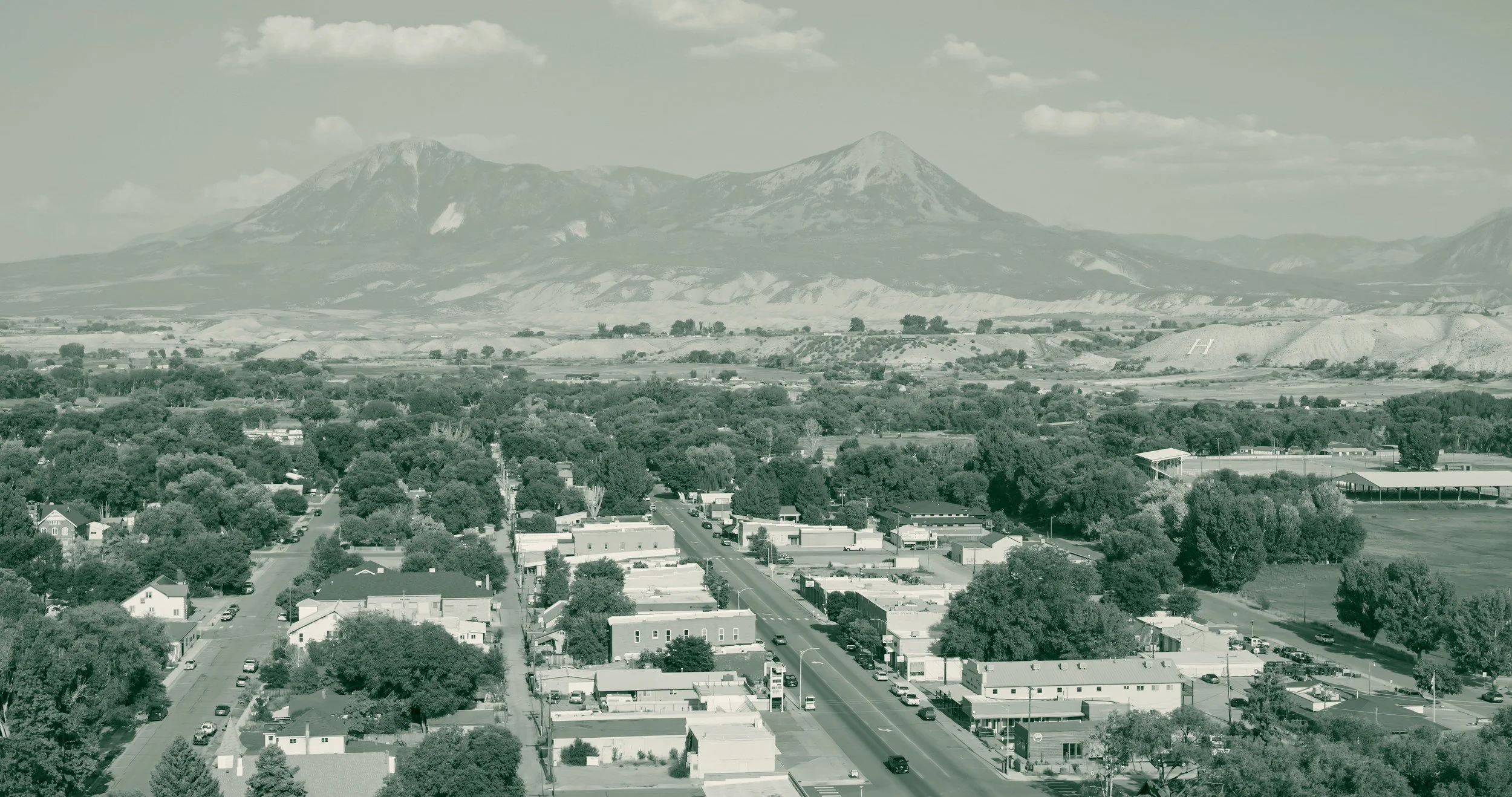 Downtown Hotchkiss, Colorado, seen from above, with low storefronts and parked cars along the main street, tree-shaded blocks and irrigated fields beyond, and a long mesa rising to two peaks behind the valley.