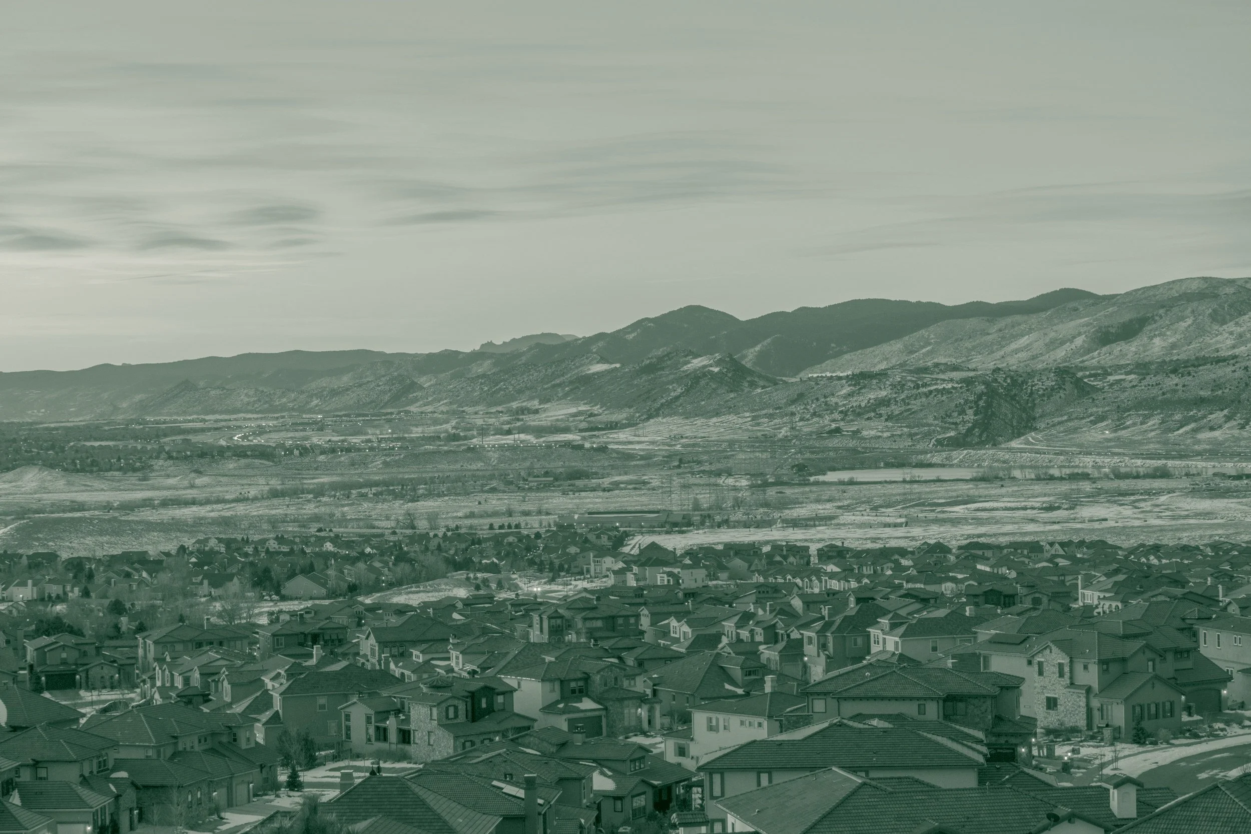 A residential neighborhood of single-family homes on the sloping side of Green Mountain in Lakewood, Colorado, at sunrise, with streets and rooftops in the foreground and the ridgeline rising behind.